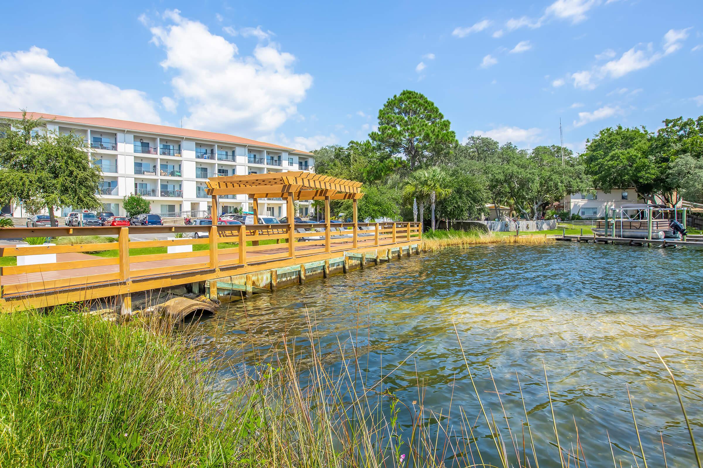 A wooden dock extending over a calm body of water, surrounded by grassy areas and trees. In the background, a three-story white building with a red roof is visible, along with parked cars. The sky is bright with a few clouds, creating a serene outdoor scene.