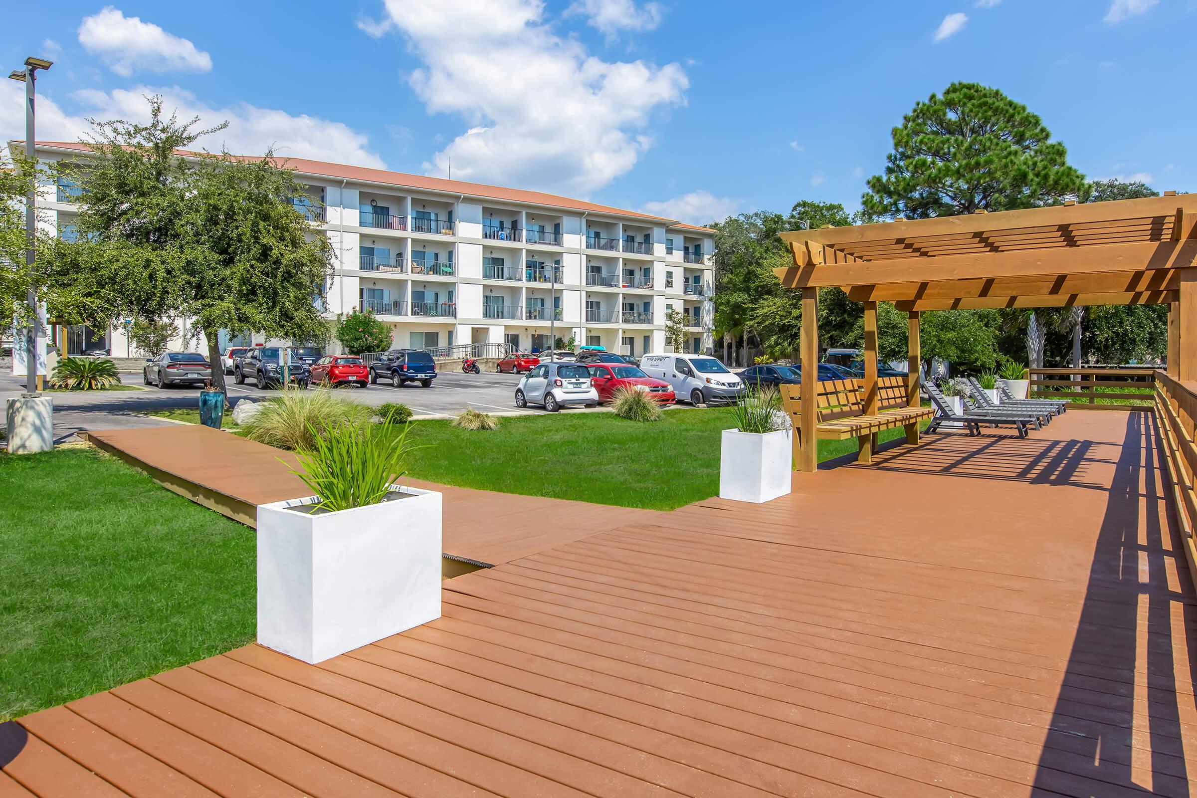 A wooden deck area featuring a pergola with benches, surrounded by green grass and ornamental plants. In the background, a multi-story residential building and parked cars are visible under a partly cloudy sky.