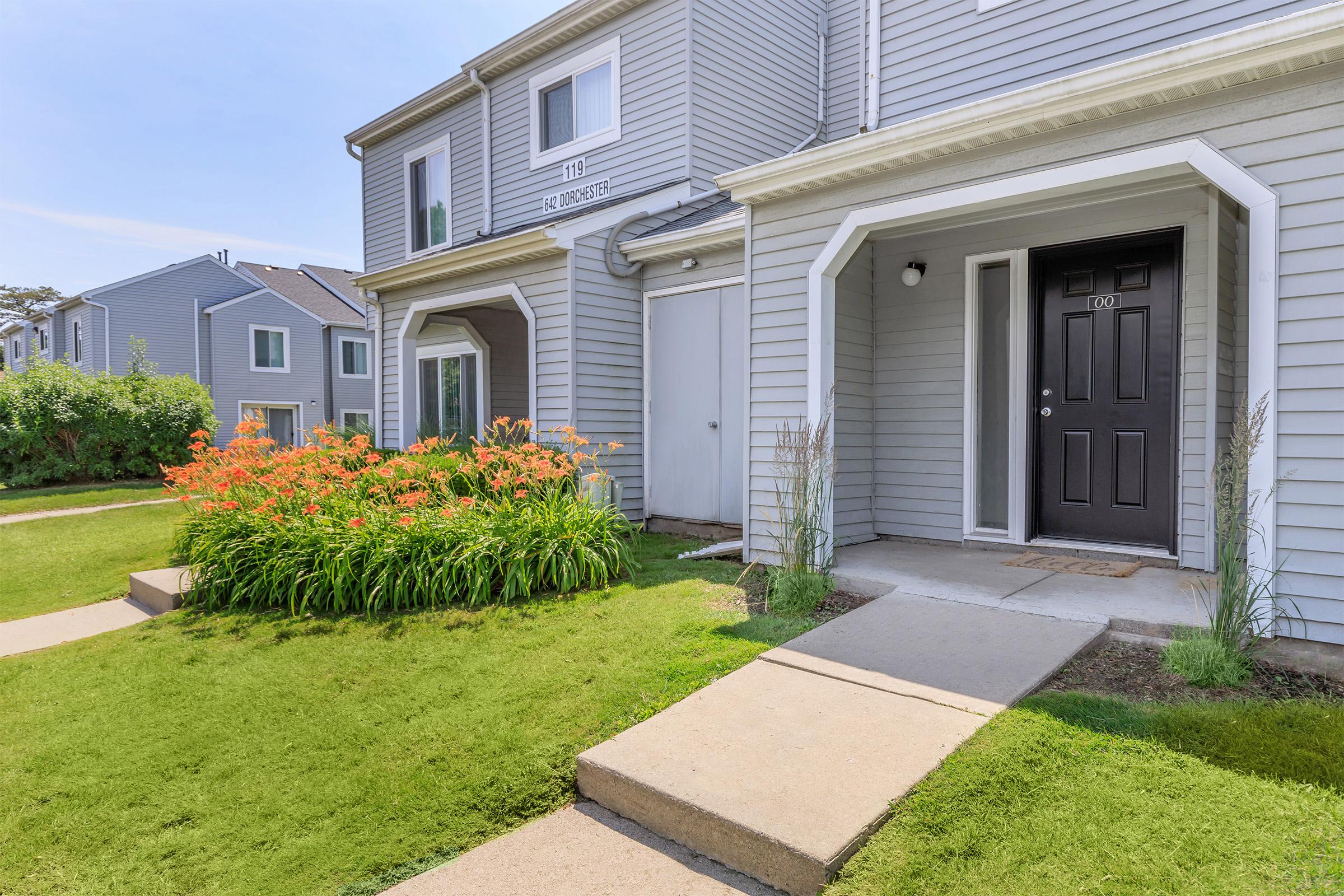 Exterior view of a residential building with two doorways. The entrances are flanked by lush green grass and colorful flower beds. The building features light gray siding and is set in a well-maintained landscape with walkways leading to each door. Bright blue sky is visible above.