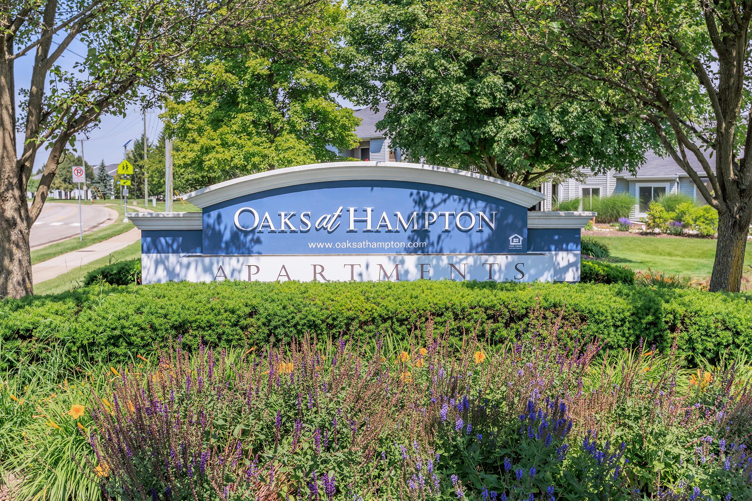 Sign for "Oaks at Hampton Apartments" displayed prominently in a landscaped area with green bushes and colorful flowers. The sign features a blue background and white lettering, set amidst trees and grass, indicating a welcoming residential community.