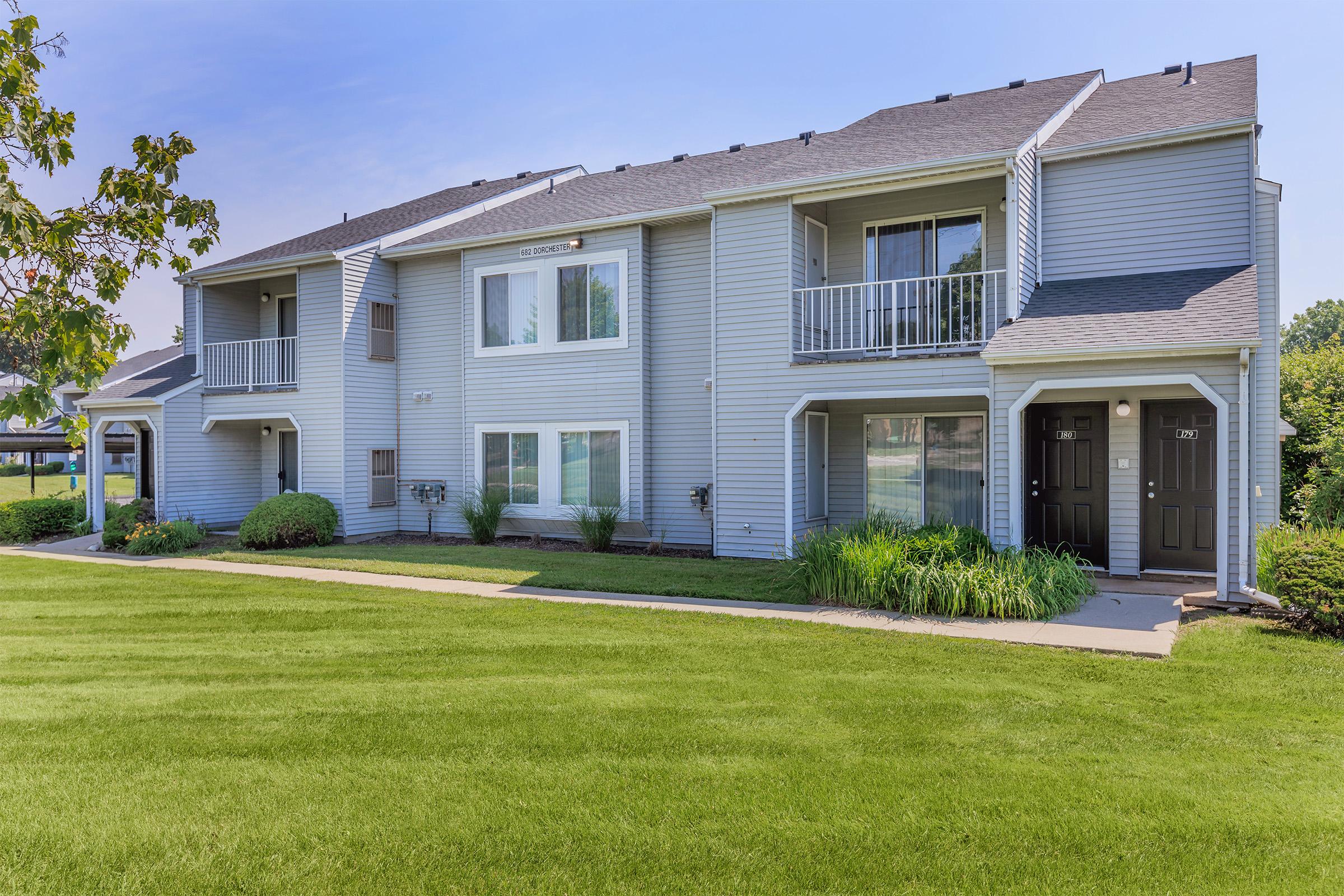 A front view of a two-story apartment building, featuring grey siding and multiple balconies. The well-maintained landscape includes green grass and shrubs, with a walkway leading up to the entrance doors. The sky is clear, suggesting a sunny day.
