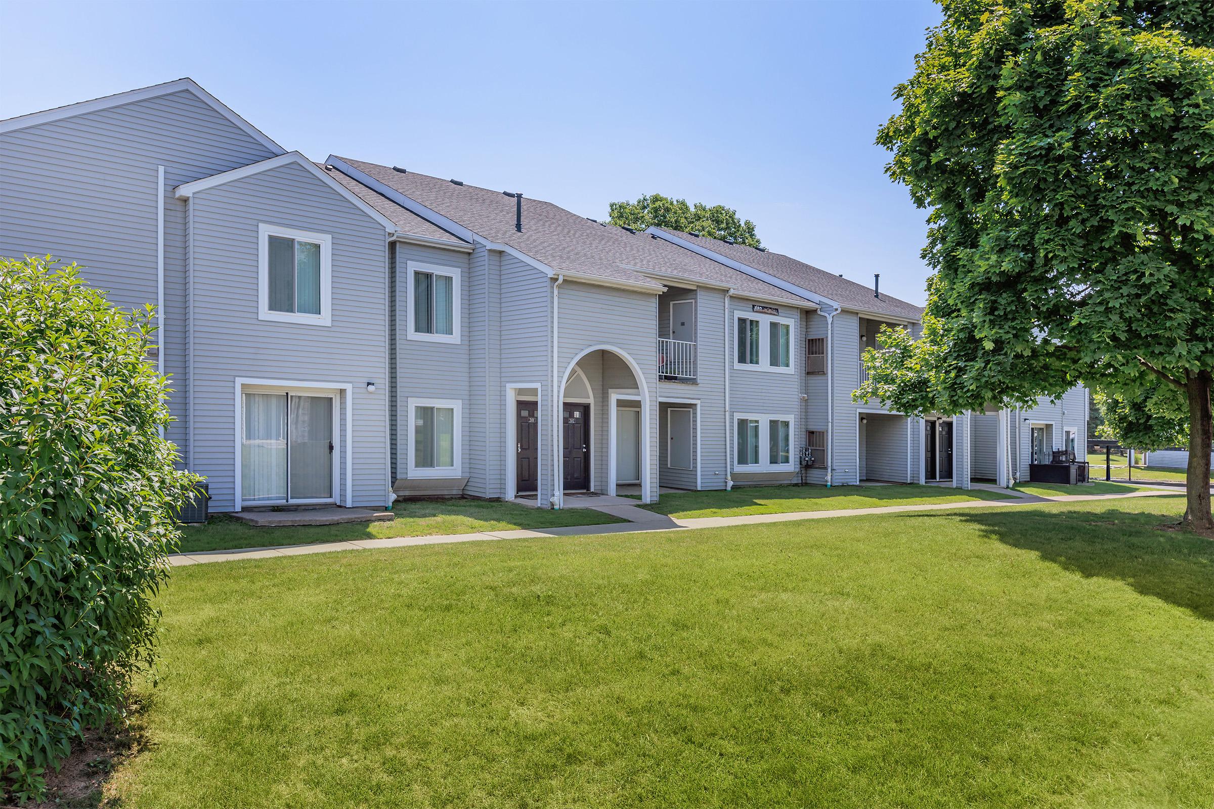A row of modern two-story apartment buildings with light gray siding. The buildings feature arched doorways and large windows, surrounded by green grass and shrubs. A tree provides shade in the foreground, and clear blue skies are visible above. The scene is bright and inviting.
