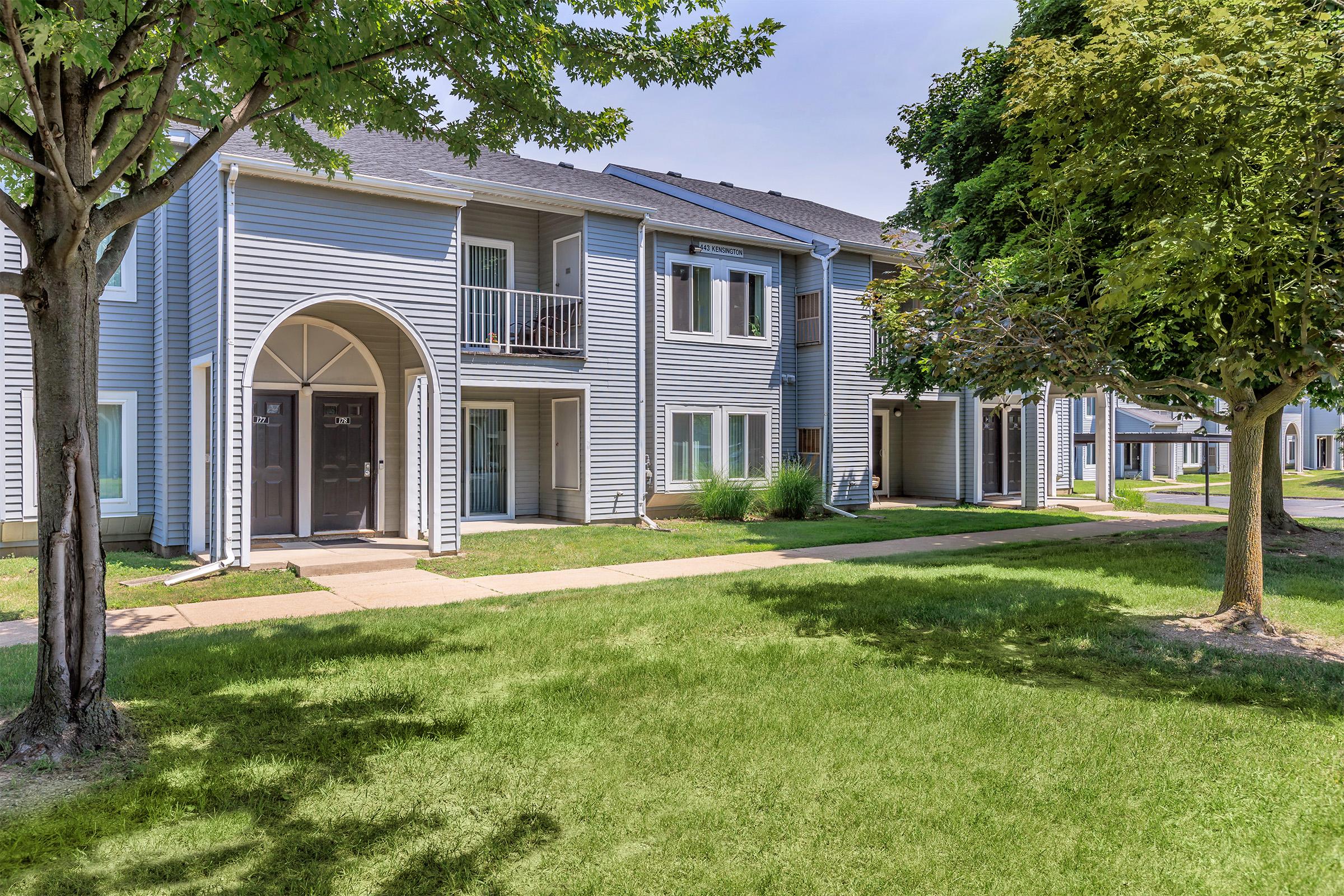 A row of modern apartment buildings featuring light blue siding, with two entrances visible. The well-maintained grassy area is bordered by trees, providing a pleasant outdoor space. The sky is clear, and the overall setting appears bright and inviting.