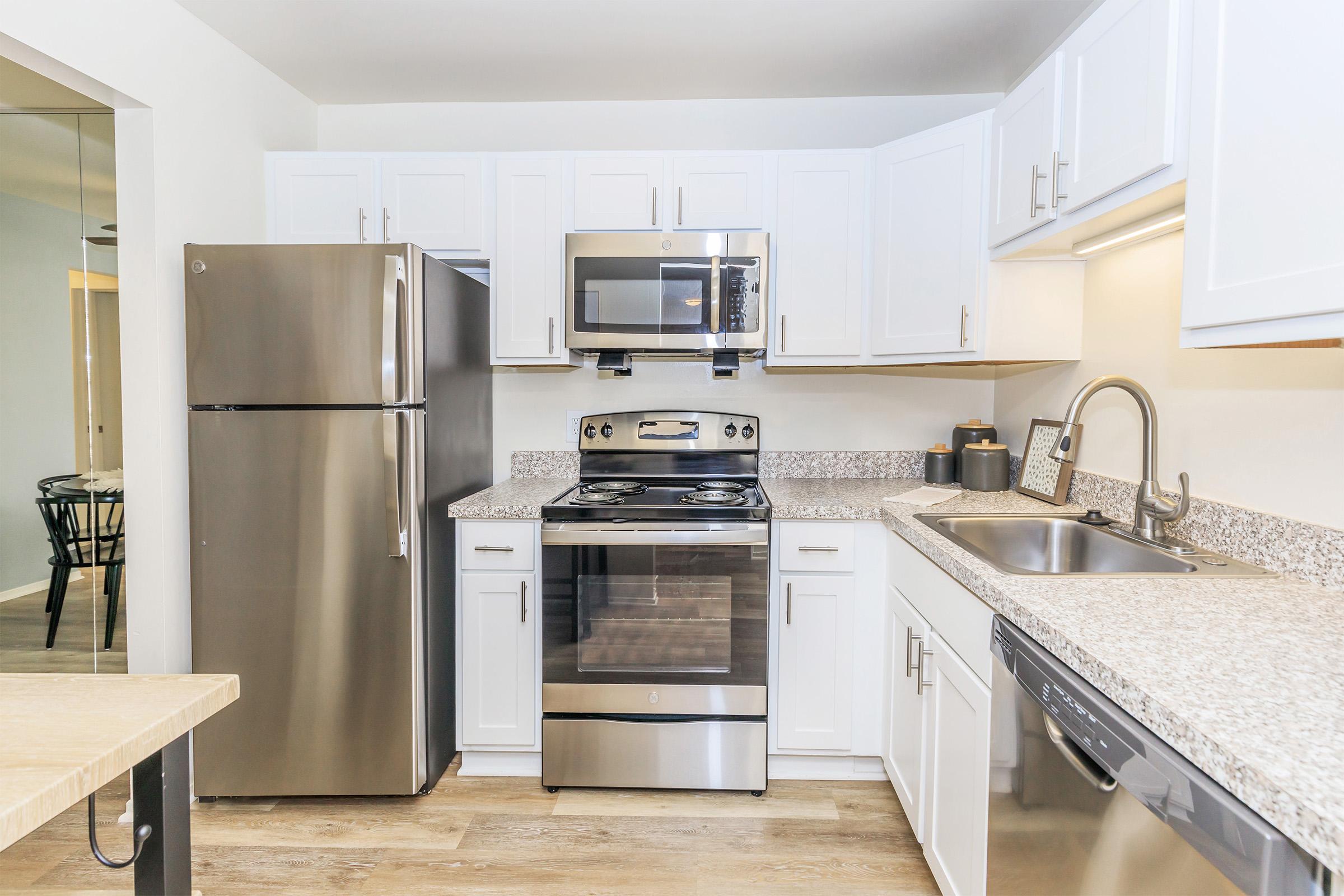 A modern kitchen featuring stainless steel appliances, including a refrigerator, oven, microwave, and dishwasher. It has white cabinetry and a light granite countertop, along with a sink and minimal decorative items. The space is well-lit and designed for efficiency.