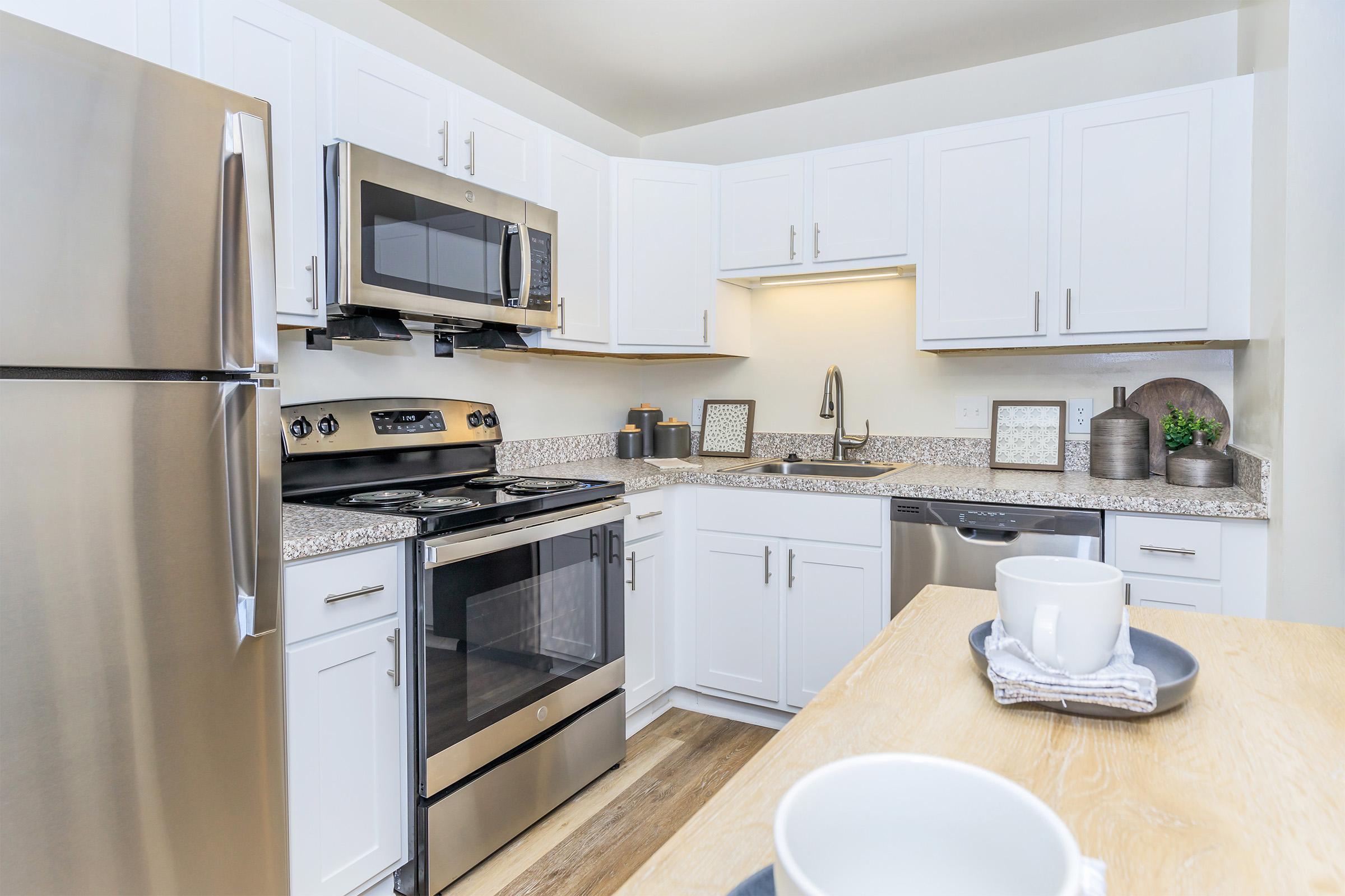 Modern kitchen featuring stainless steel appliances, including a refrigerator, oven, and microwave. The white cabinetry and granite countertops create a sleek look. There's a sink with a modern faucet, and a wooden table with two coffee cups in the foreground, adding a cozy touch to the space.