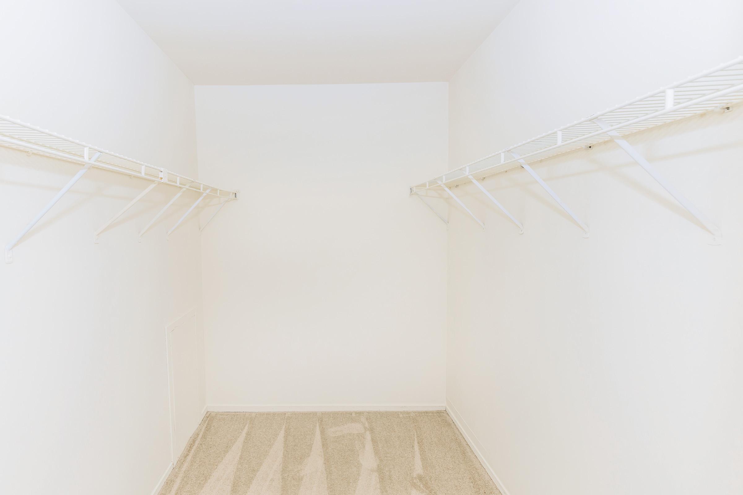A spacious, empty walk-in closet featuring two white wire shelves on opposite walls, with bare beige carpet flooring and plain white walls. The closet is well-lit and shows no items or storage, creating a minimalistic appearance.