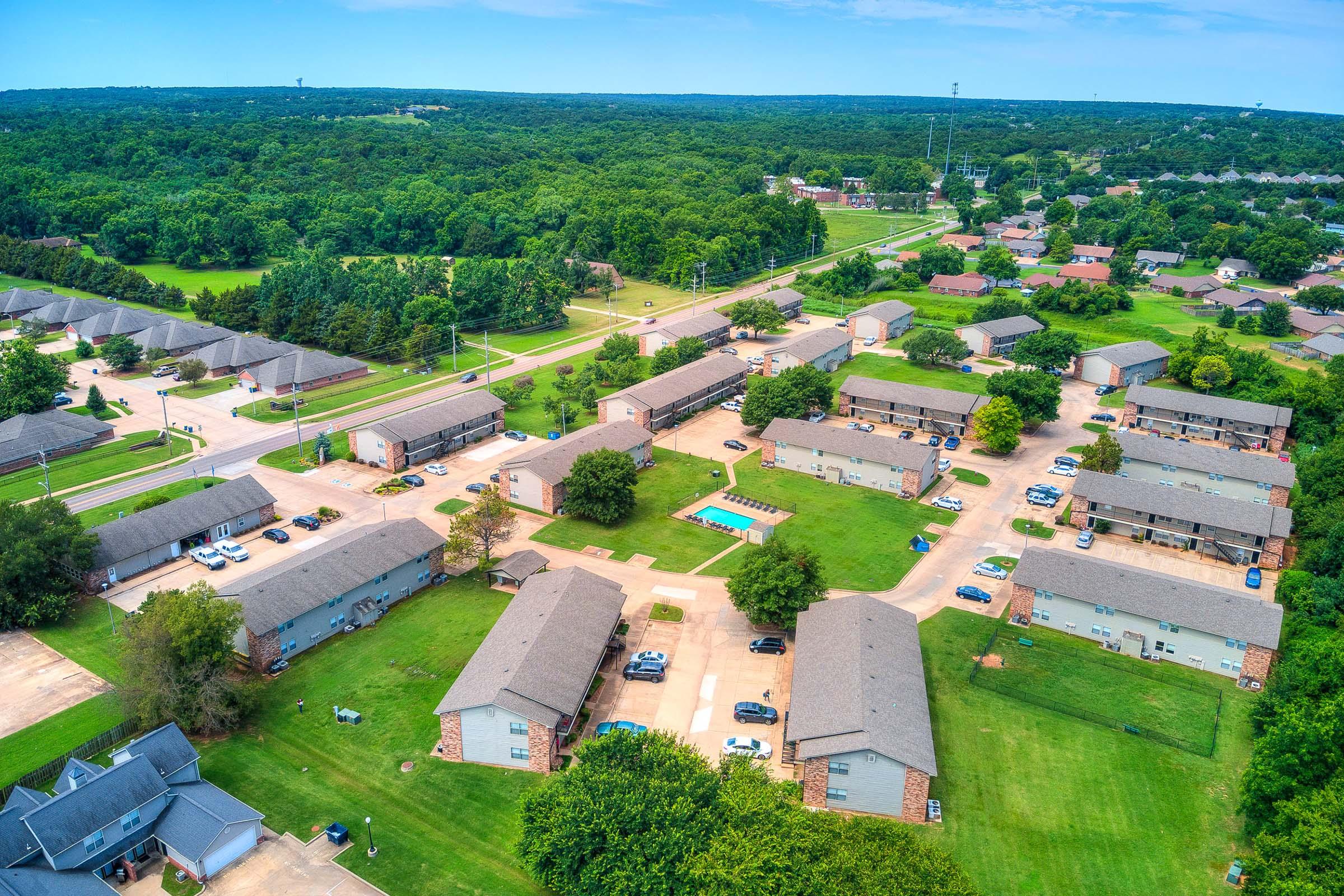 Aerial view of a residential neighborhood featuring multiple low-rise apartment buildings arranged around green lawns and a swimming pool. Tree-covered areas and roads with parked cars are visible, showcasing a suburban environment with a mix of construction and natural landscapes.