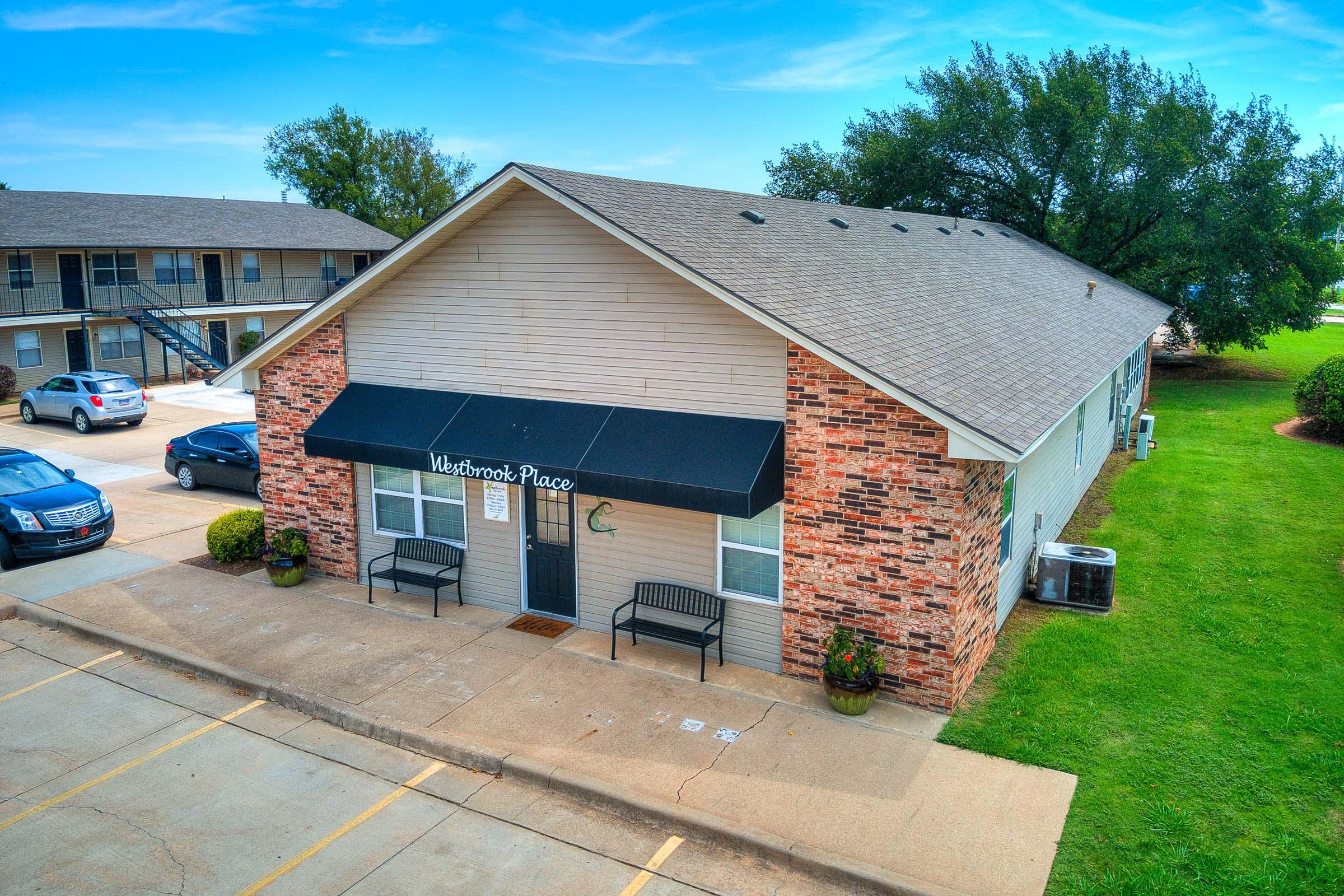 A single-story building with a brick facade and a dark awning that reads "Waterbrook Place." It features benches outside, surrounded by manicured lawns. In the background, there are additional buildings and parked cars. The sky is clear with a few clouds.