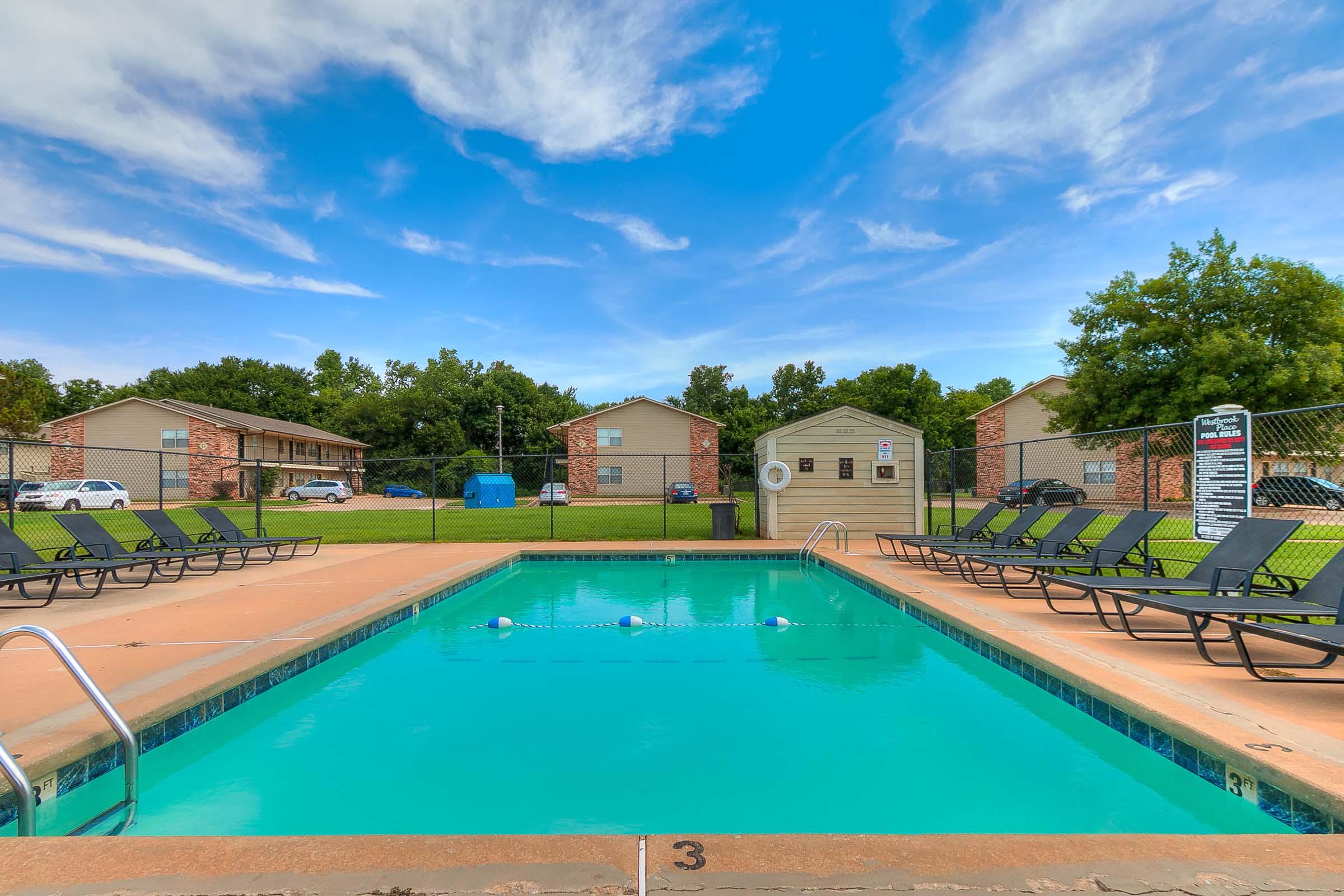A swimming pool surrounded by lounge chairs, set against a backdrop of apartment buildings and green trees under a blue sky with fluffy clouds. The pool area is inviting, with clear water and a safety sign nearby.