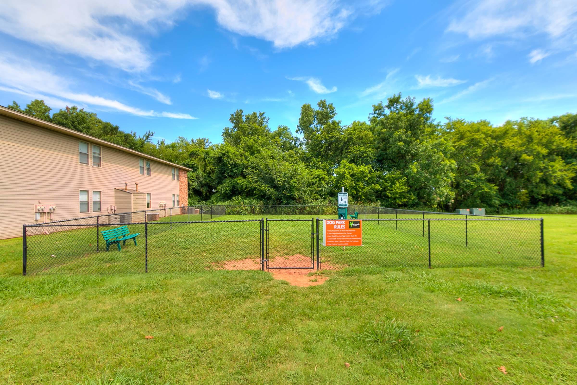 Fenced area in a green grassy space with a sign indicating a dog park. A bench is visible, and trees line the background under a blue sky with scattered clouds. The park is designed for pets to play and socialize.