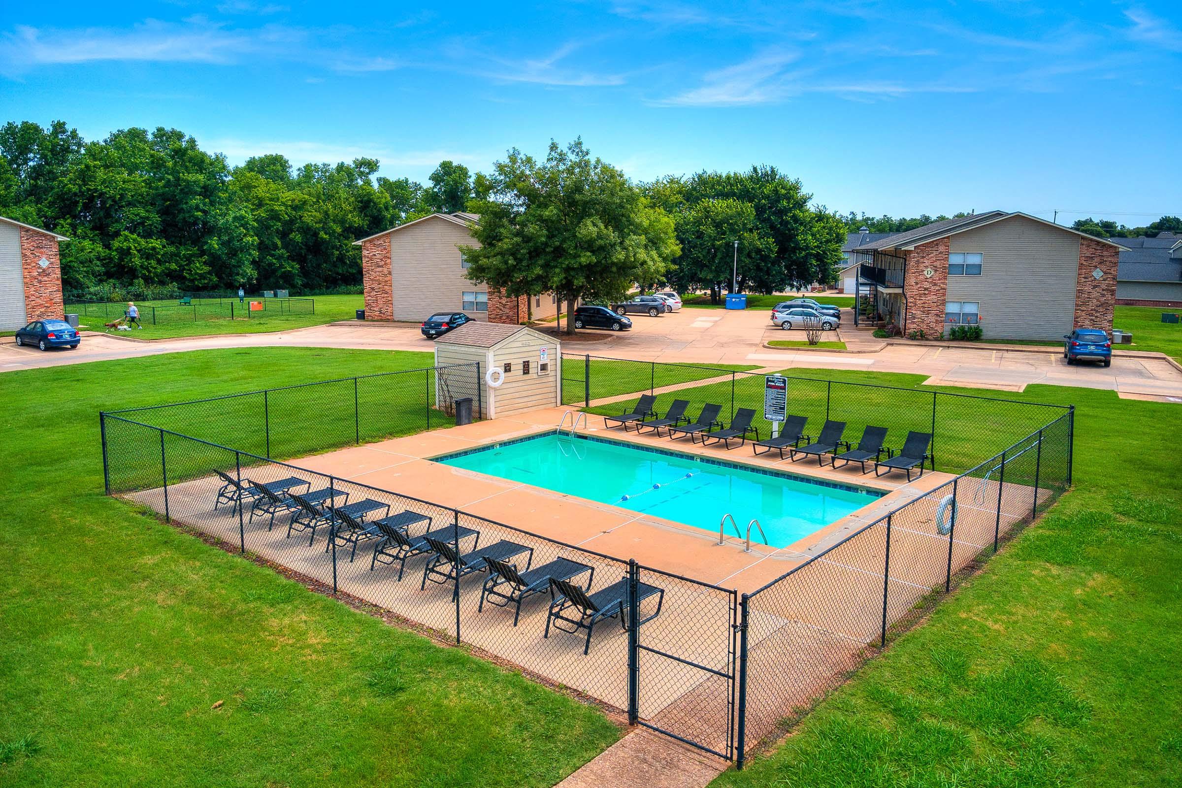 An outdoor swimming pool surrounded by a fence, with lounge chairs lined up on the patio. In the background, several brick apartment buildings are visible along with cars parked in a lot. The area is lush with green grass and trees under a clear blue sky.