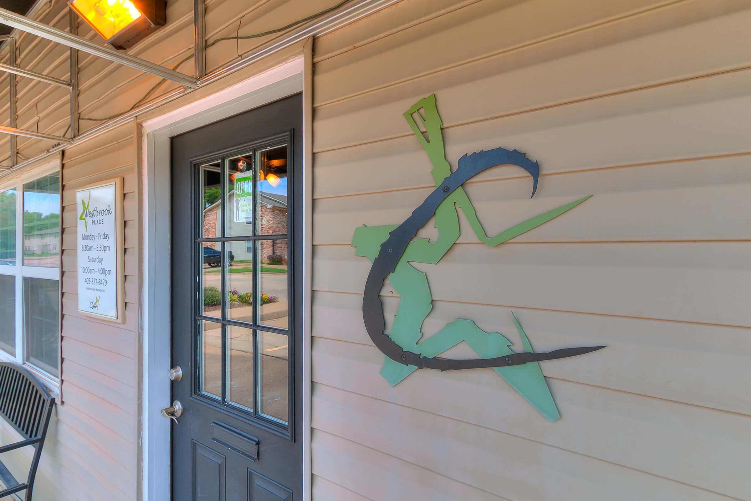A decorative sign featuring a stylized representation of a person with a curved object, positioned next to a door. The building's exterior is light-colored with a dark door, and there is a sign nearby with business hours. A bench is also visible in front of the entrance.