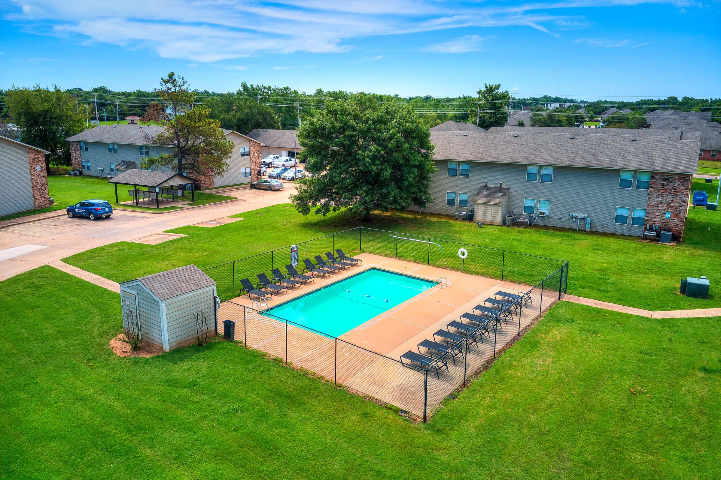 Aerial view of a residential area featuring a swimming pool surrounded by a fenced deck with lounge chairs. Nearby are two gray apartment buildings set in a grassy courtyard, with trees and parked cars in the background under a clear blue sky.