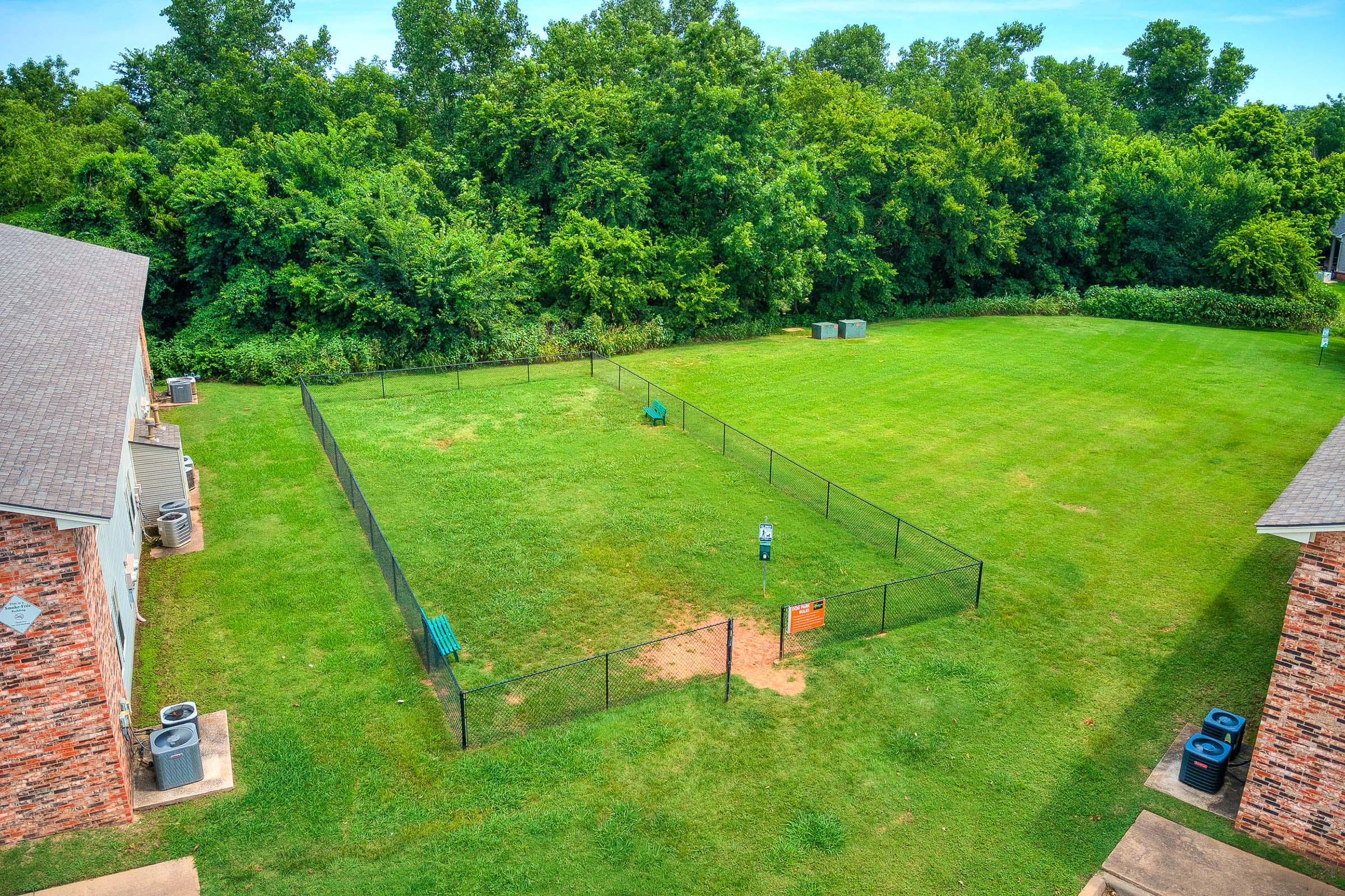 Aerial view of a grassy area enclosed by a black fence, featuring two benches and a sign. The space is bordered by thick greenery and includes two air conditioning units visible near the buildings on either side. The sky is clear with a few clouds.