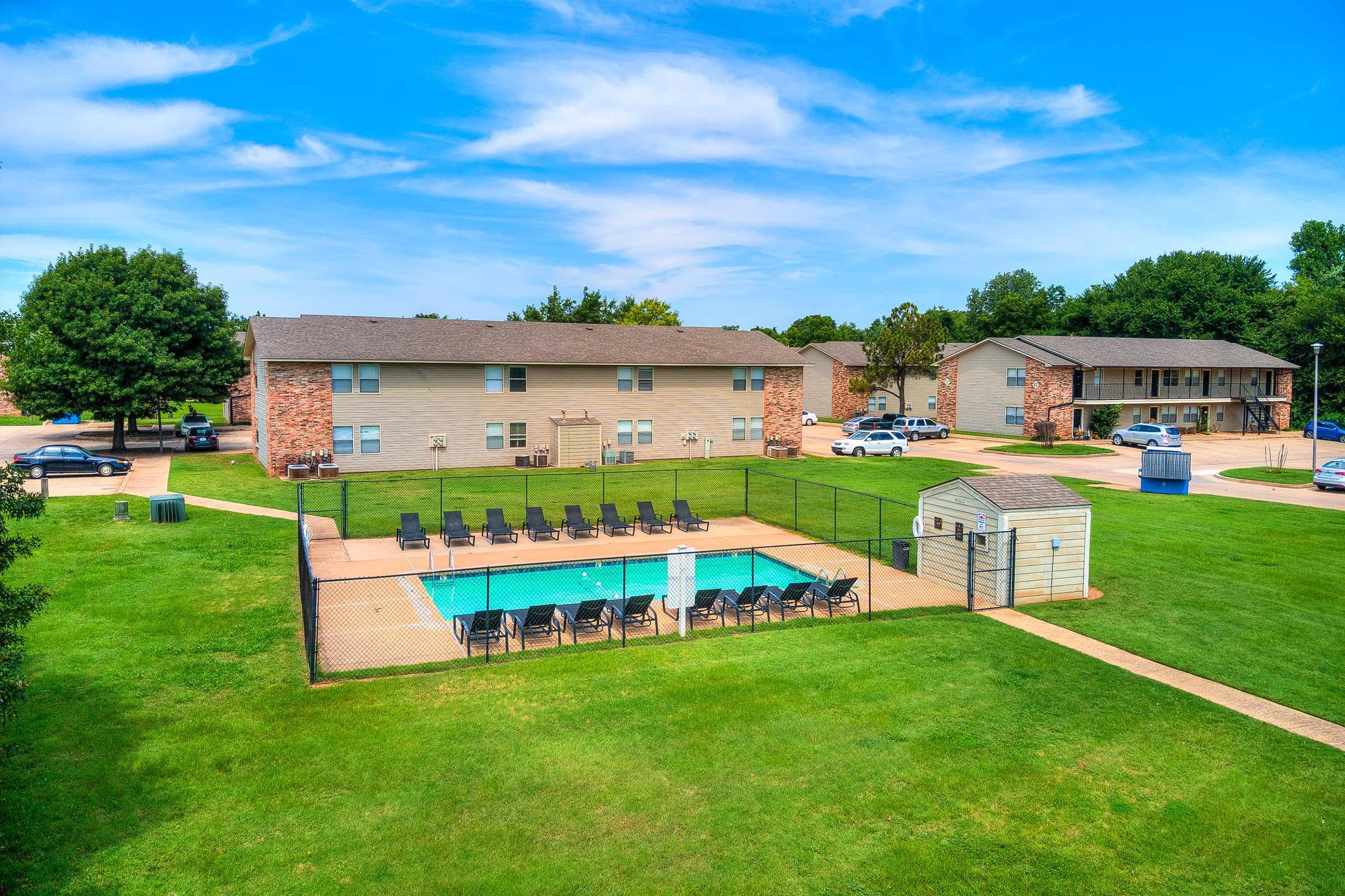 Aerial view of an apartment complex featuring a swimming pool surrounded by lounge chairs. The green lawn and trees complement the three buildings in the background. Clear blue skies enhance the inviting atmosphere of the outdoor space.