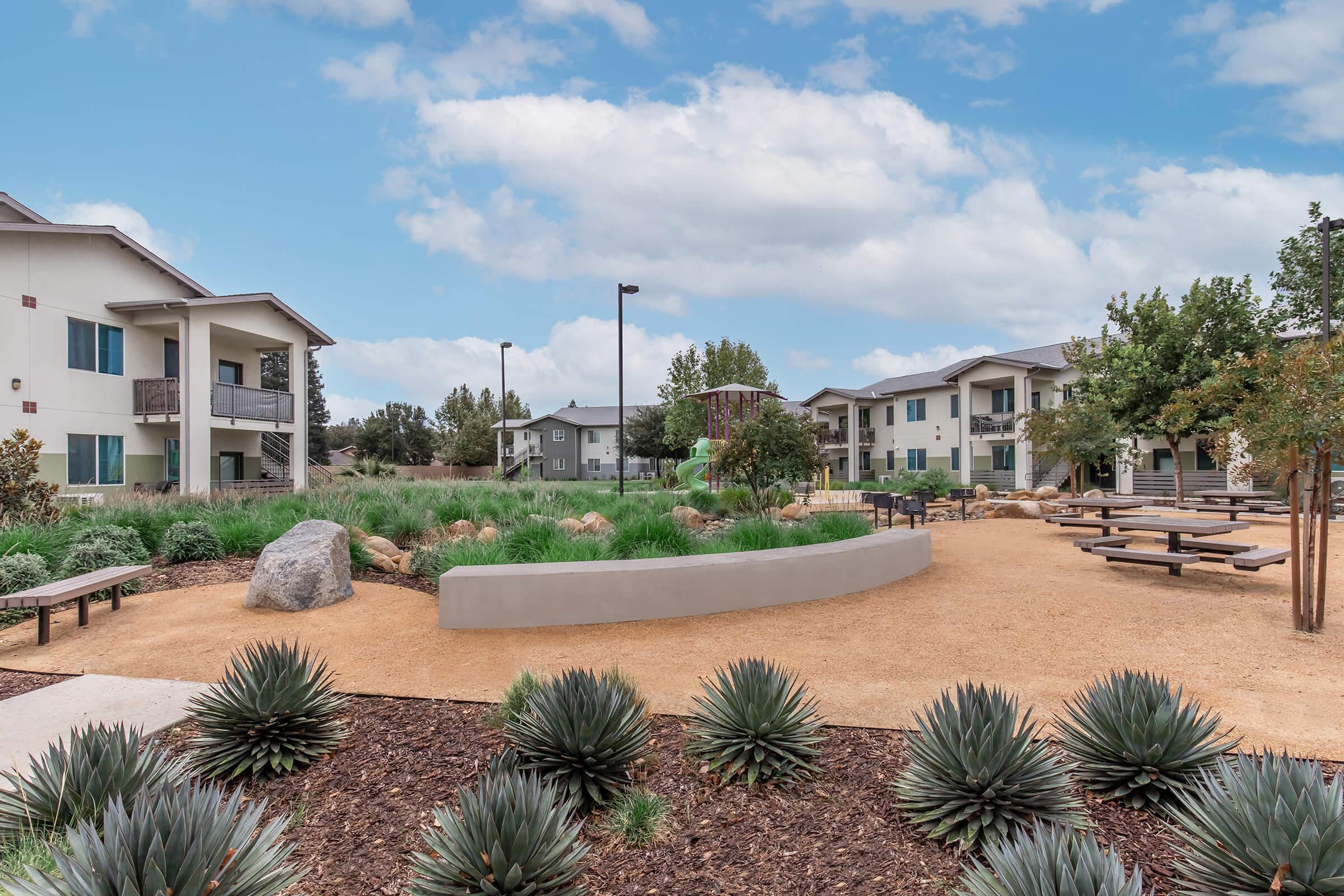 A landscaped courtyard featuring a mix of gravel and grass, with modern apartment buildings in the background. There are picnic tables, a curved seating area, boulders, and shrubs, under a partly cloudy sky, creating a welcoming outdoor community space.