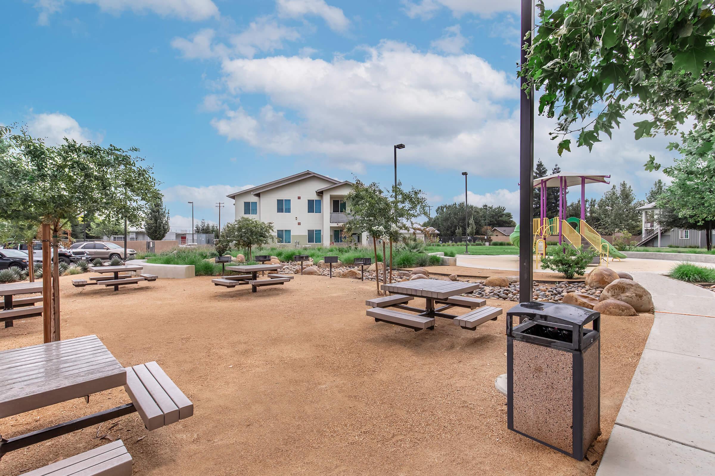 A spacious outdoor park area featuring picnic tables, gravel pathways, and a playground in the background. Nearby are trees and landscaped areas with rocks. In the distance, there are residential buildings and parking spaces visible under a partly cloudy sky.