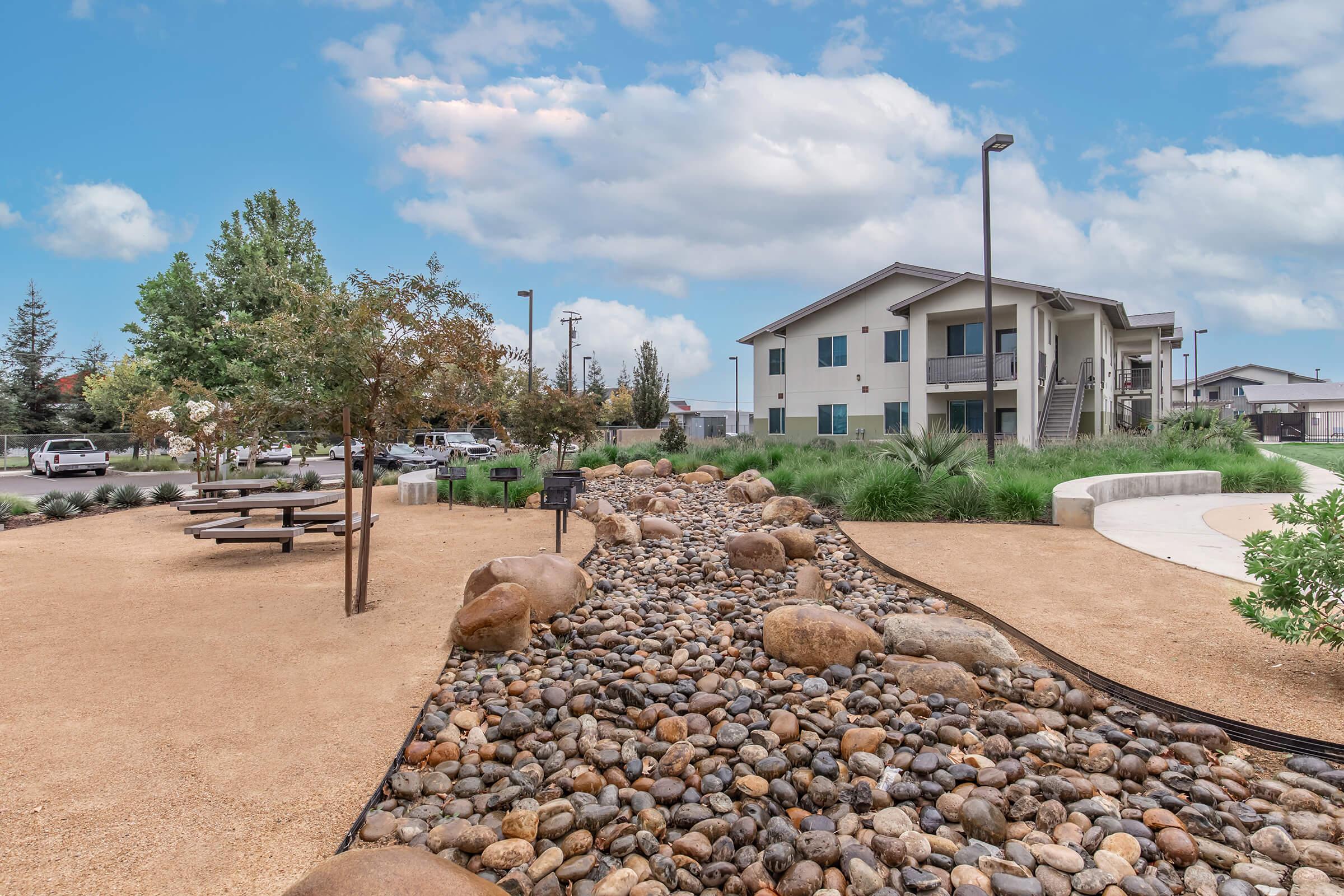 A landscaped area featuring a dry riverbed lined with smooth rocks. There are trees and shrubs in the background, along with picnic tables. In the distance, a multi-story residential building is visible under a partly cloudy sky. The overall scene conveys a calm, outdoor environment.