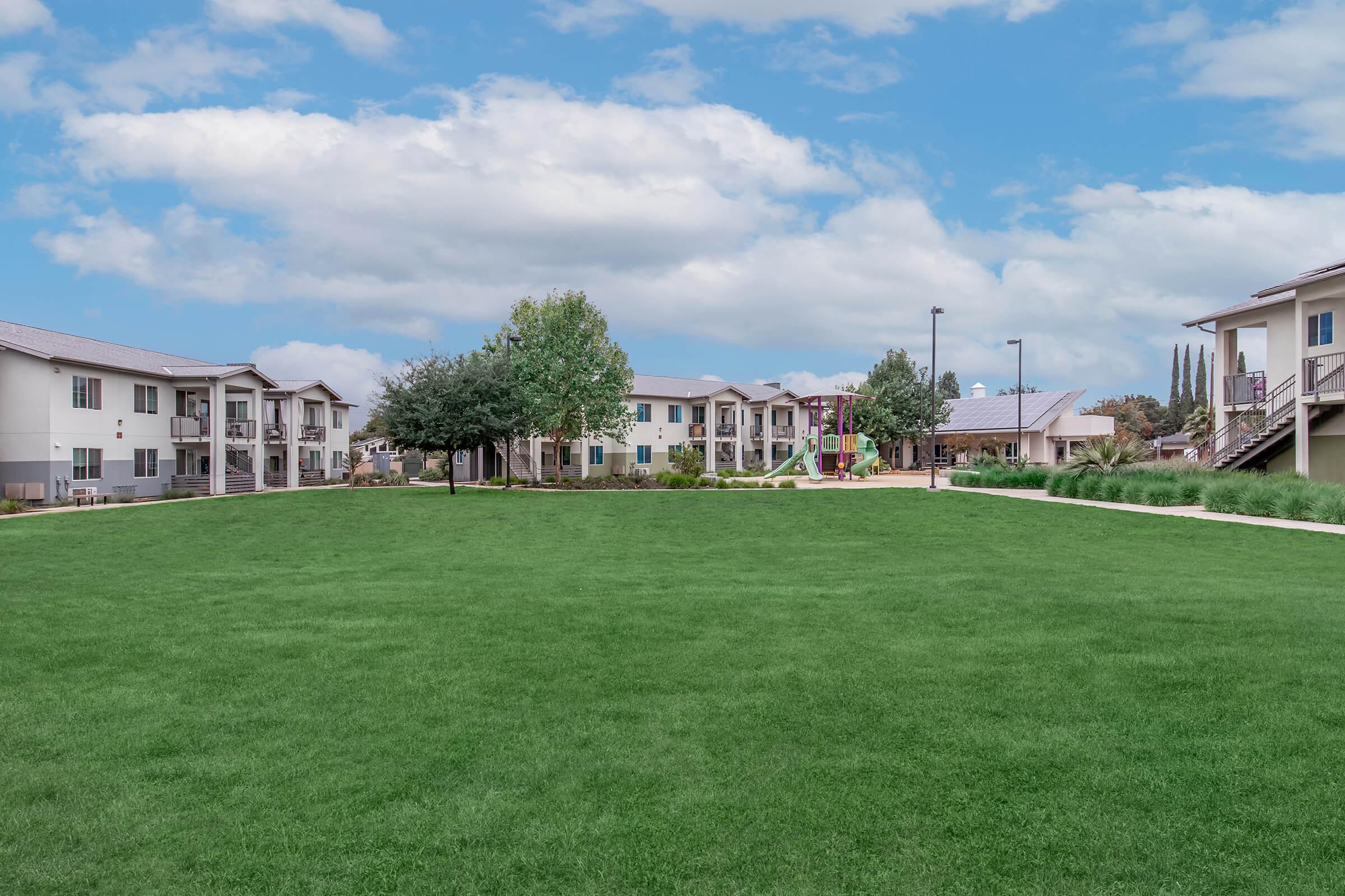 A well-maintained green lawn surrounded by residential buildings. The scene features multiple two-story apartment complexes with balconies, trees, and a playground in the background under a blue sky with clouds. The area appears inviting and spacious, suitable for outdoor activities.