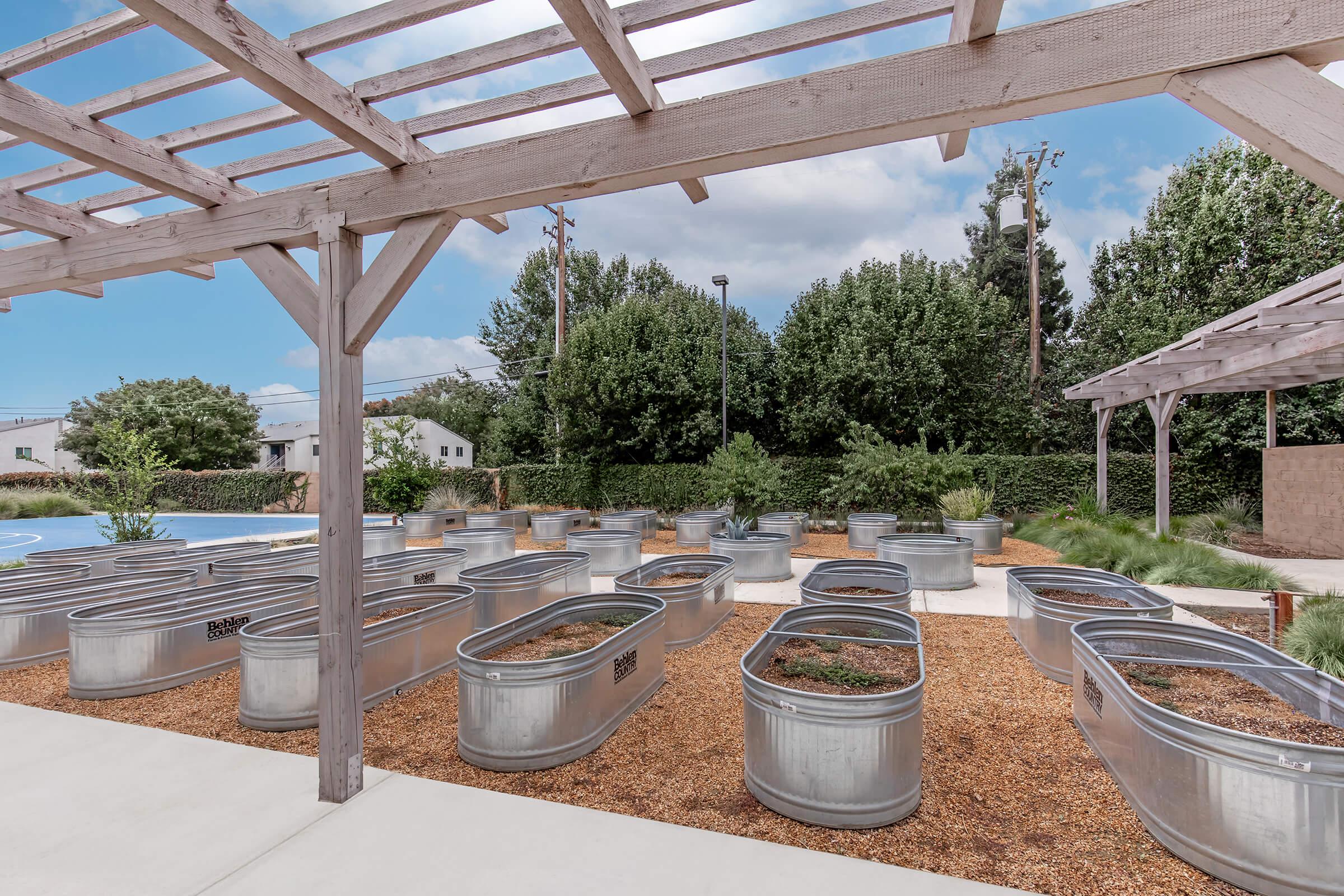 A landscaped area featuring multiple metal planters arranged in neat rows, surrounded by gravel. A wooden pergola provides shade, with a clear blue sky and green trees in the background. The setting appears to be designed for gardening or community use.