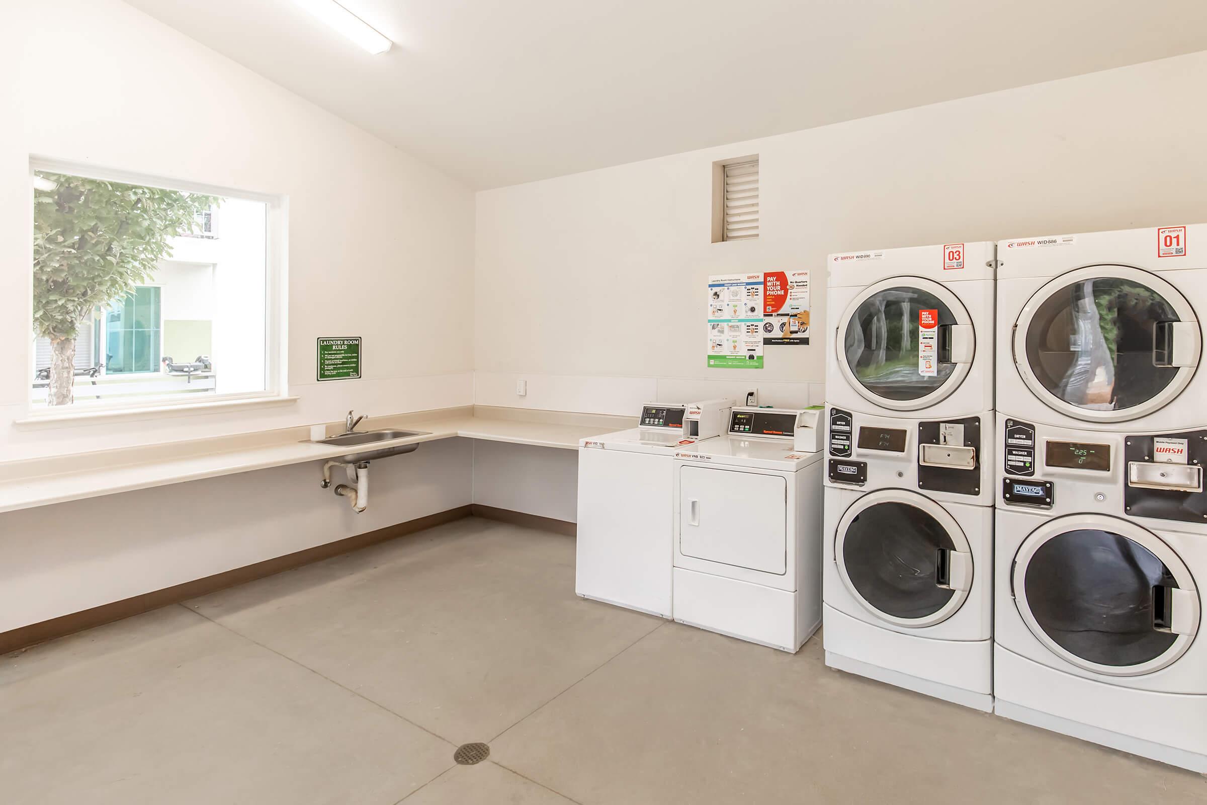 A clean, modern laundry room featuring multiple washing machines and dryers. There is a countertop space for folding clothes and a small sink. A window allows natural light to enter, and a bulletin board with laundry instructions is visible on the wall. The floor is concrete with a drain in the center.
