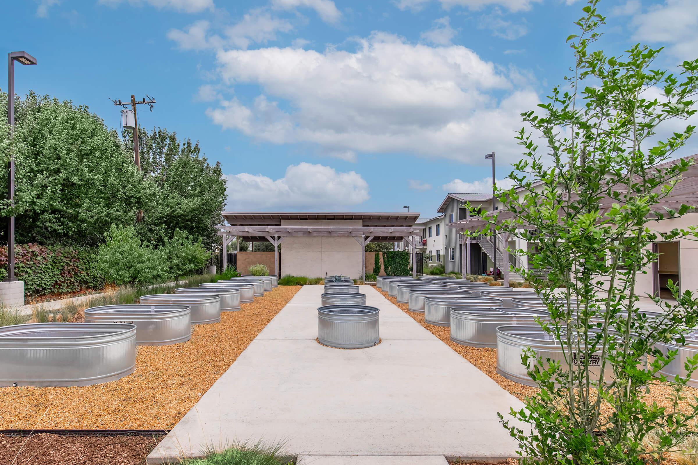 A scenic outdoor area featuring a row of metal raised garden beds surrounded by gravel and landscaping. In the background, there is a wooden pavilion and buildings partially obscured by greenery under a bright, partly cloudy sky.