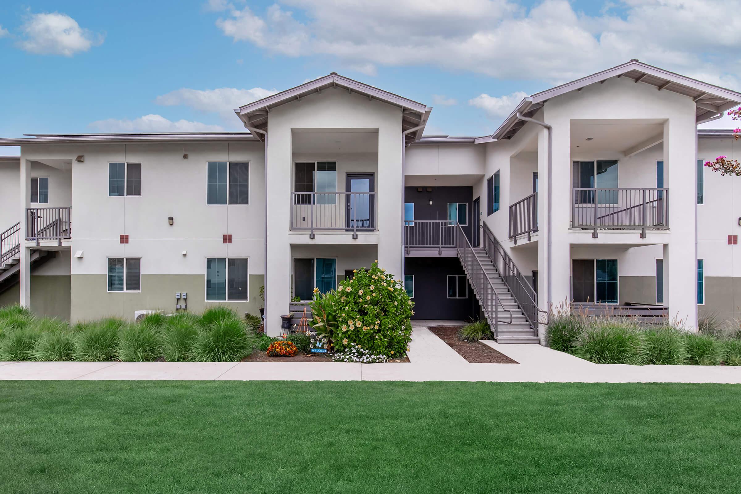A modern apartment building with two levels, featuring multiple balconies and a landscaped front yard with green grass and flower beds. The building has a neutral color scheme, large windows, and a walkway leading to the entrance. The sky is partly cloudy, adding to the pleasant setting.