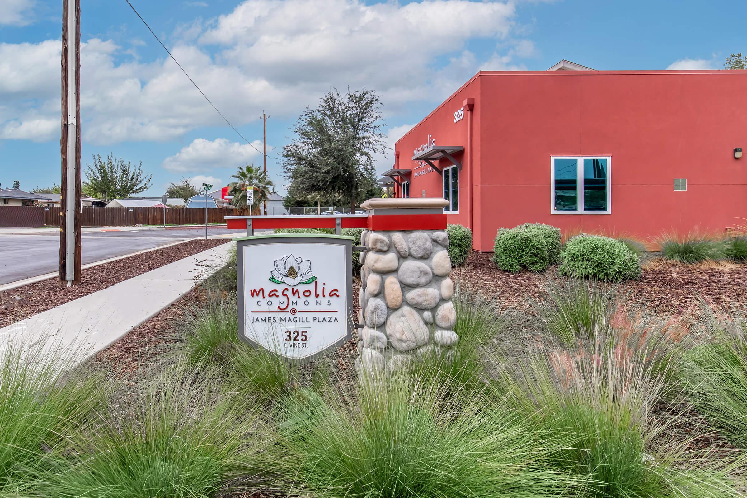 A sign for Magnolia Commons located at 325 James Magill Plaza, featuring a decorative post with stone elements. The setting includes low shrubs and grasses, with a bright building in the background and a blue sky.