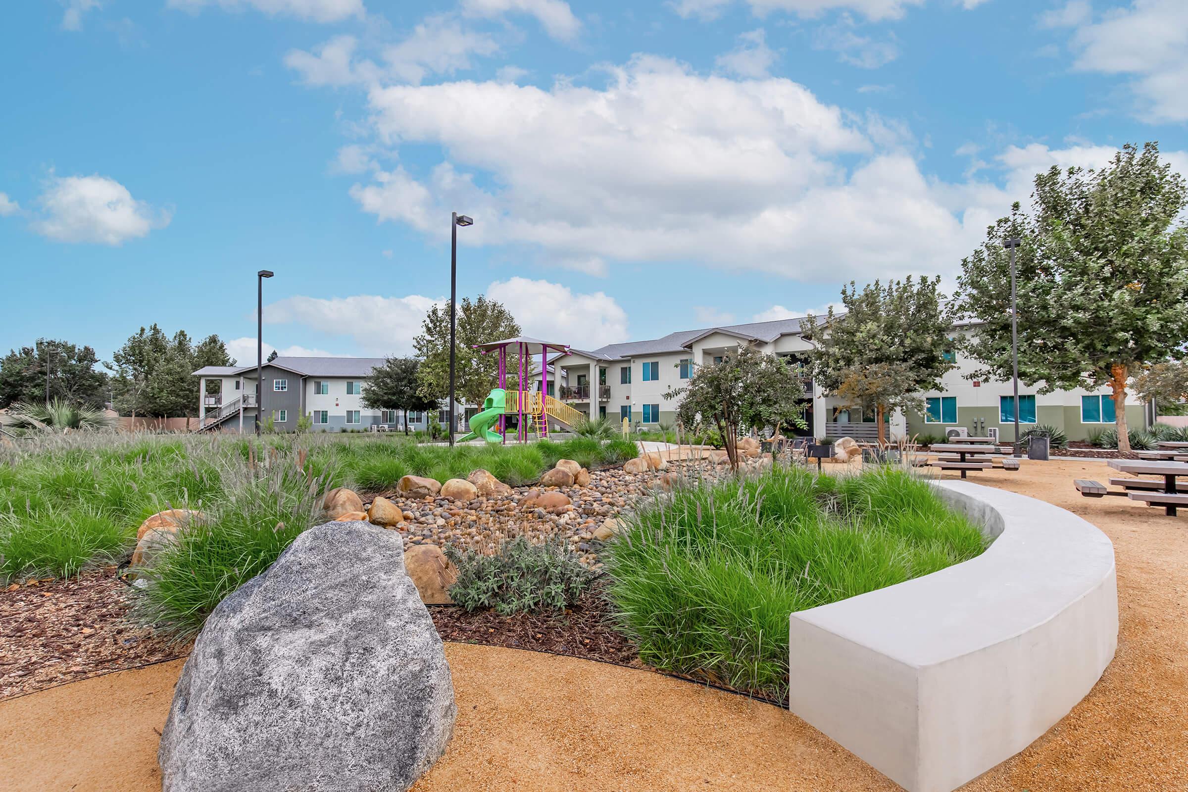 A landscaped park area featuring a curved stone bench, a large rock, and colorful play equipment in the background. Lush green grass, shrubs, and trees are visible, along with a clear blue sky and a few buildings in the distance. Ideal for recreation and relaxation.