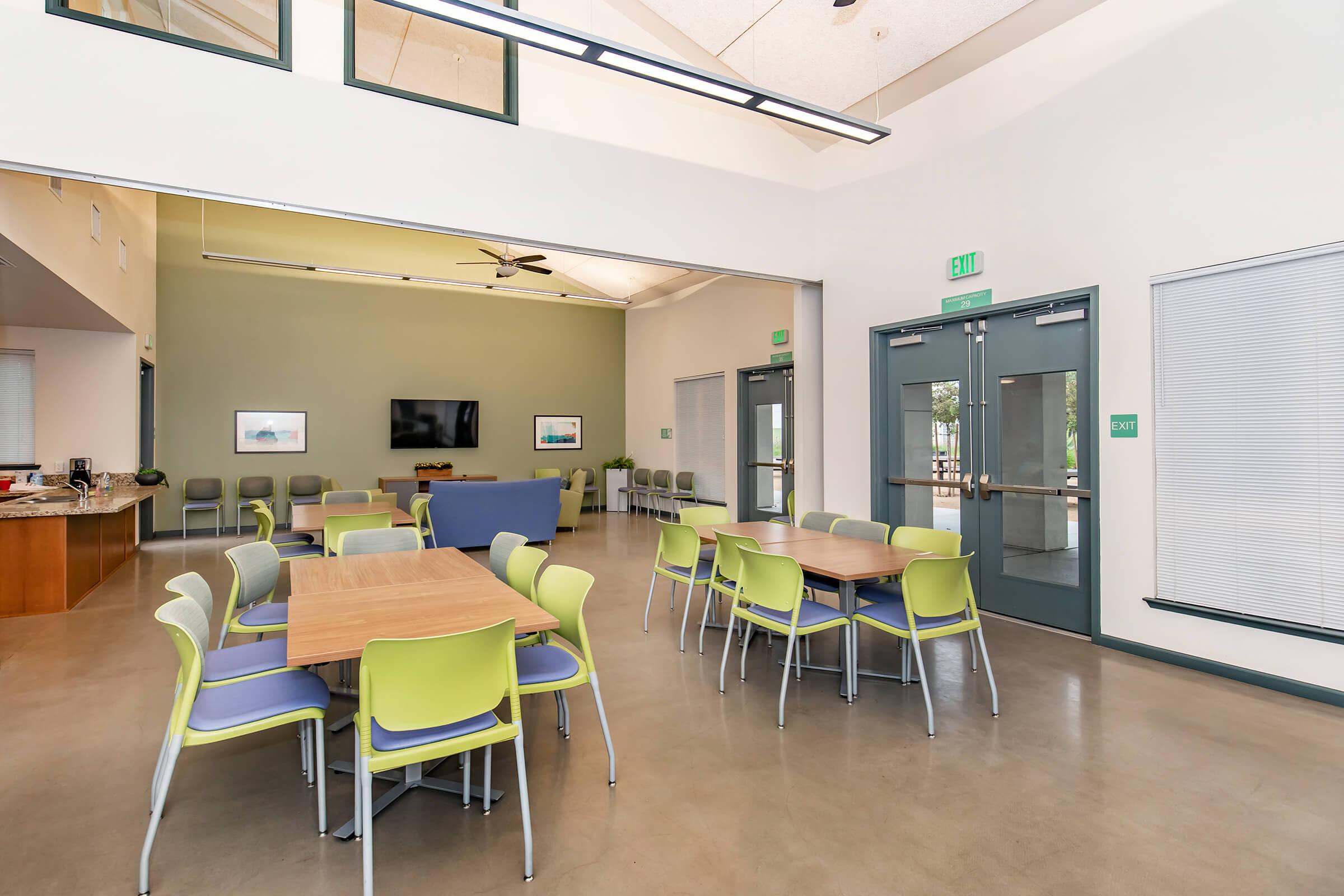 Interior view of a spacious multi-purpose room featuring several round tables with green and blue chairs. There is a kitchenette area in the background, a large television mounted on the wall, and large windows allowing natural light. The ceiling has exposed beams, enhancing the modern design.