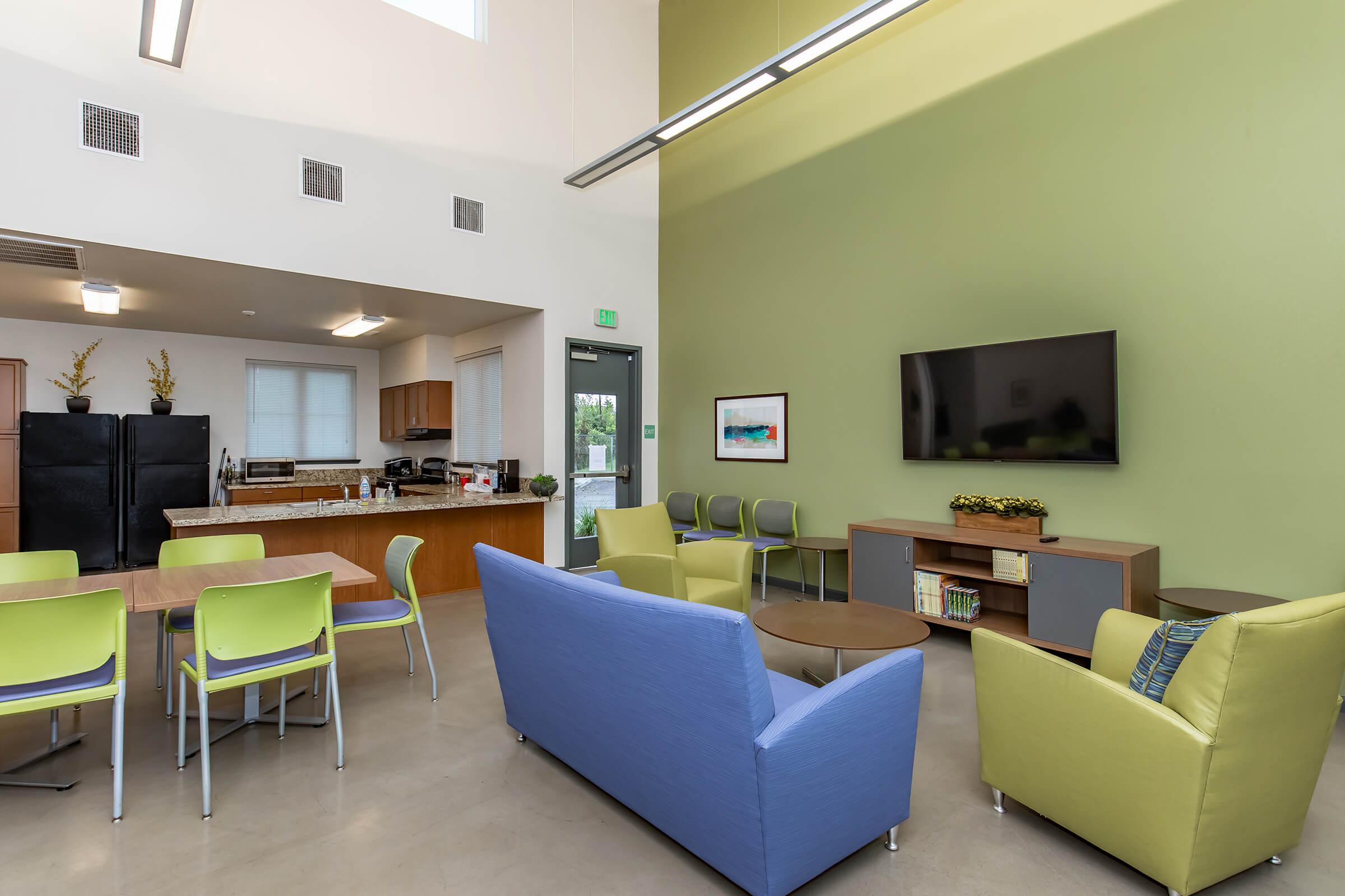 A modern communal space featuring a kitchen area with black appliances, a dining table with green chairs, and a lounge area with blue and green couches. Large windows let in natural light, and a flat-screen TV is mounted on the wall, enhancing the inviting atmosphere.