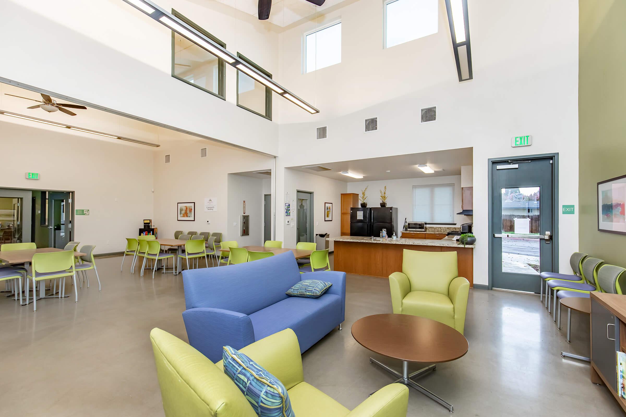 A spacious interior of a community room featuring modern furnishings. There are blue and green chairs and sofas around a circular coffee table. Large windows allow natural light in, and there is a kitchenette area visible in the background. The walls are painted in neutral tones, creating a welcoming atmosphere.