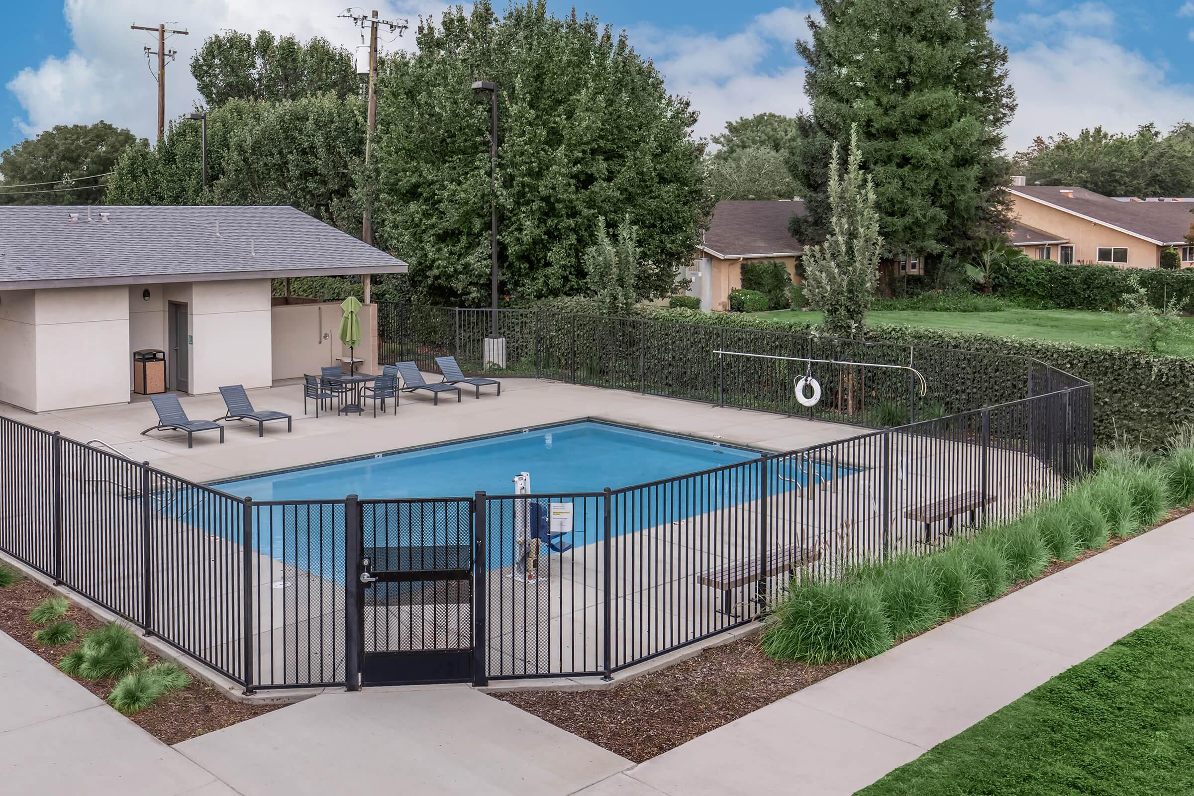 A view of a residential swimming pool area featuring a rectangular pool surrounded by lounge chairs. The pool is enclosed by a black fence, with a gate near the entrance. Nearby, there are trees and green shrubs, along with some light cloud coverage in the sky.