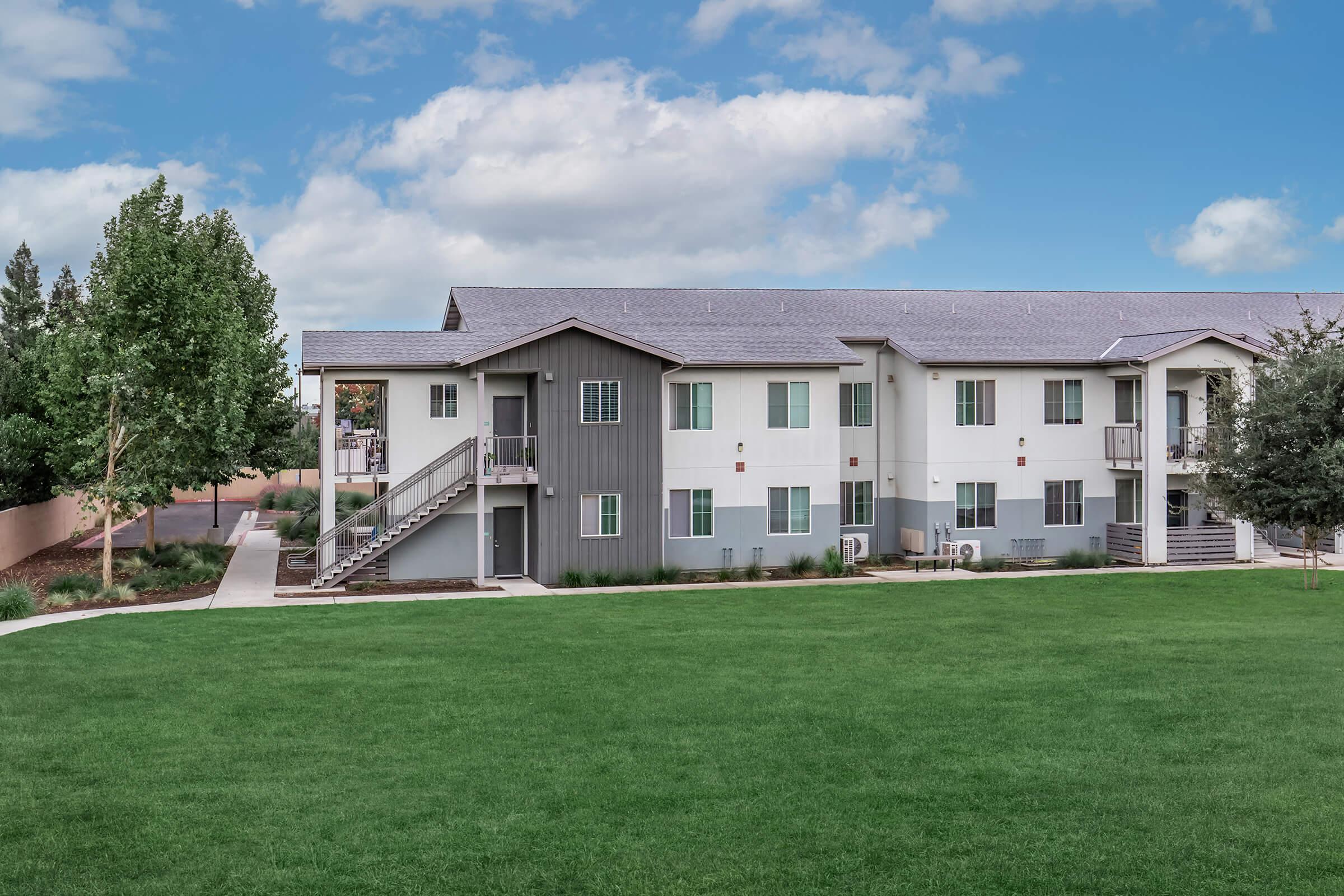 Modern multi-unit residential building with a mix of gray and white siding, surrounded by landscaped green lawn and trees. A sidewalk leads to the entrance, and the building features multiple staircases and balconies. The sky above is partly cloudy.