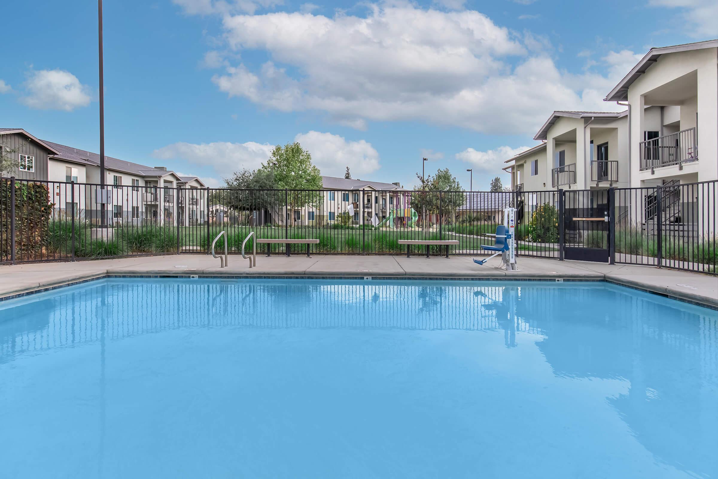 Swimming pool surrounded by a black fence with green benches nearby. In the background, modern residential buildings and greenery under a partly cloudy blue sky. The pool has clear blue water, reflecting the surroundings.