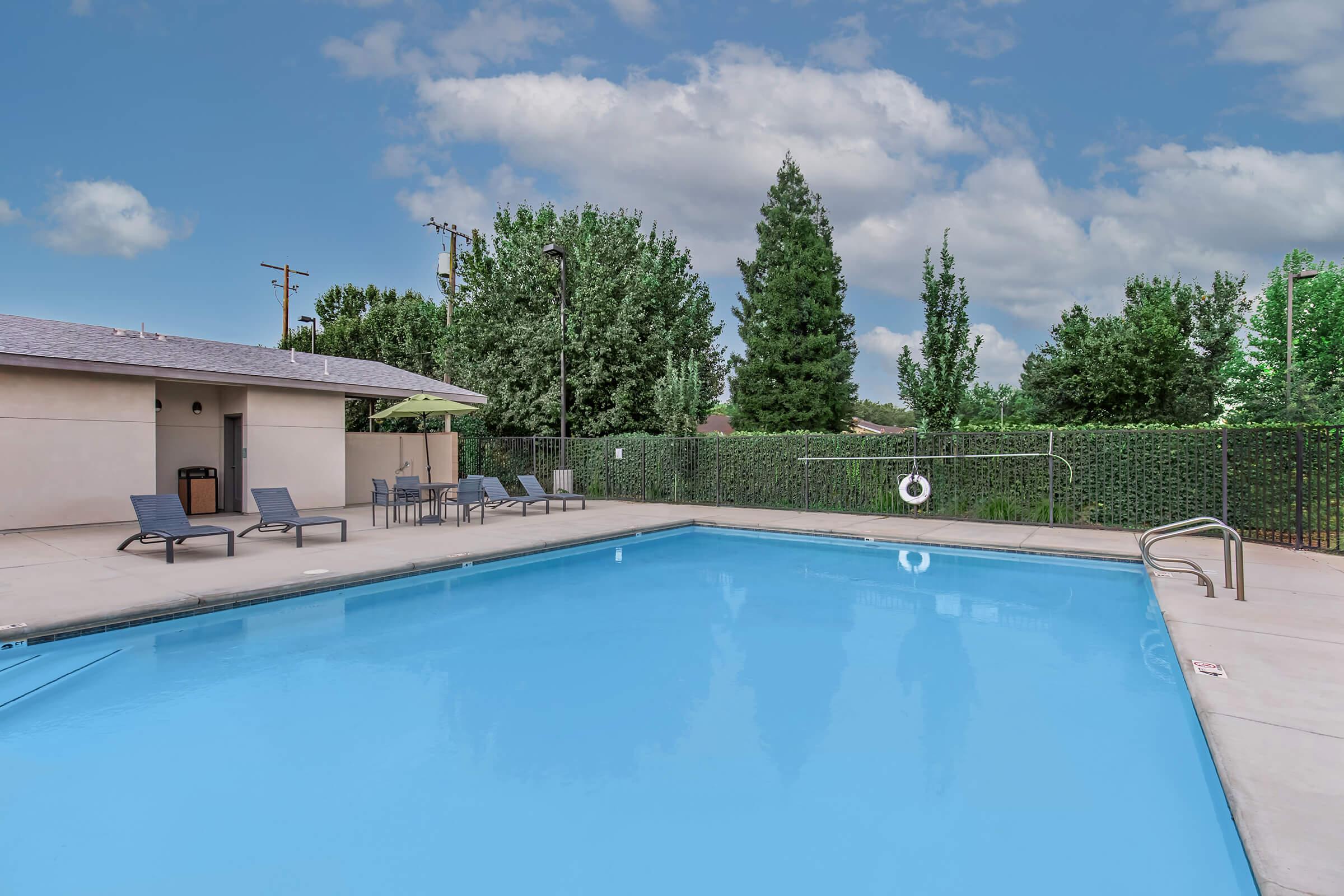 A clear blue swimming pool surrounded by a concrete deck and several lounge chairs. In the background, there are tall trees and a privacy fence, with a cloudy sky overhead. A small building is visible to the left, providing shade and amenities.