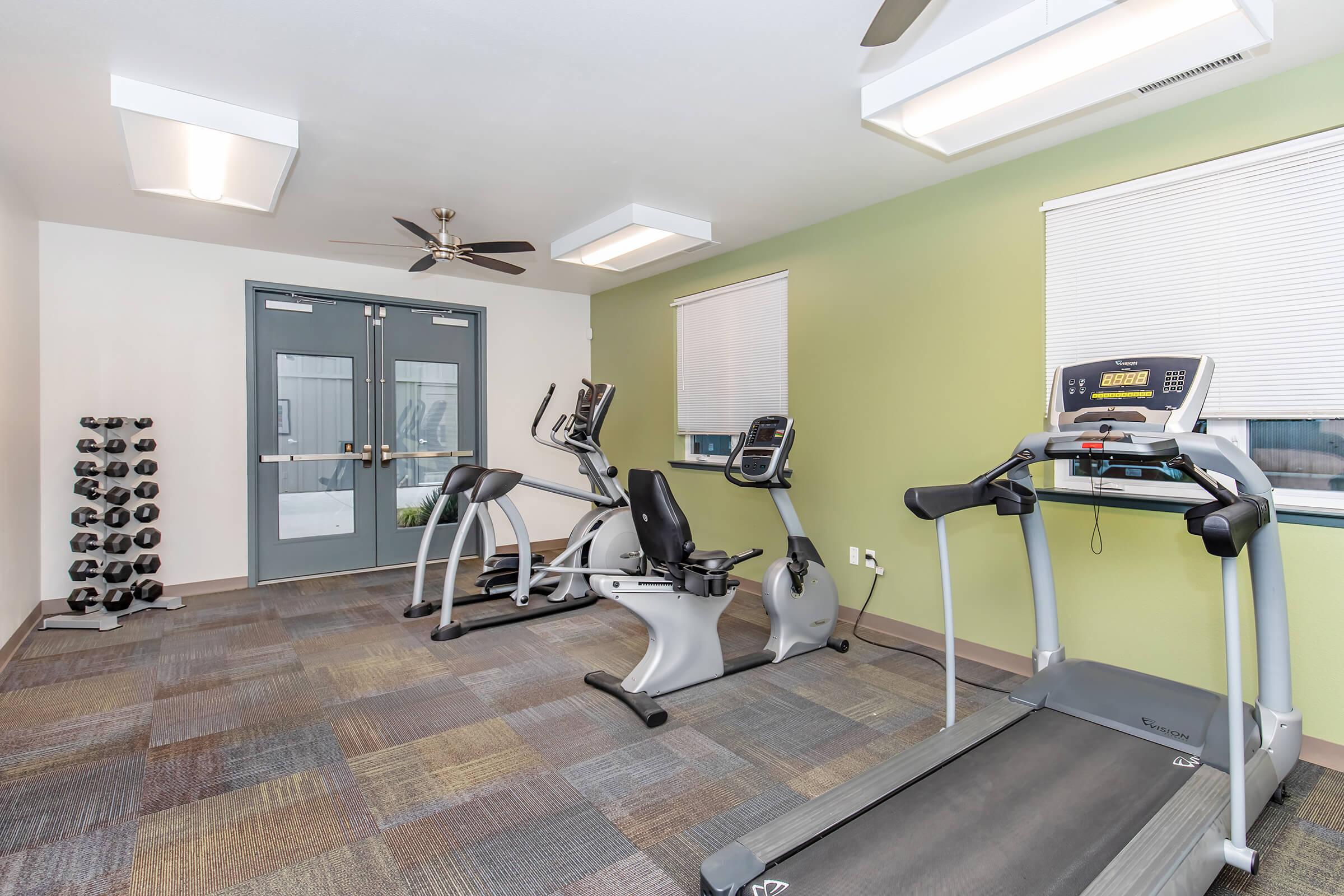 A well-lit gym area featuring exercise equipment. There are two cardio machines (an elliptical and a treadmill) and a set of dumbbells on a rack. The room has light green walls, large windows with blinds, and a ceiling fan, creating a modern and inviting workout space.