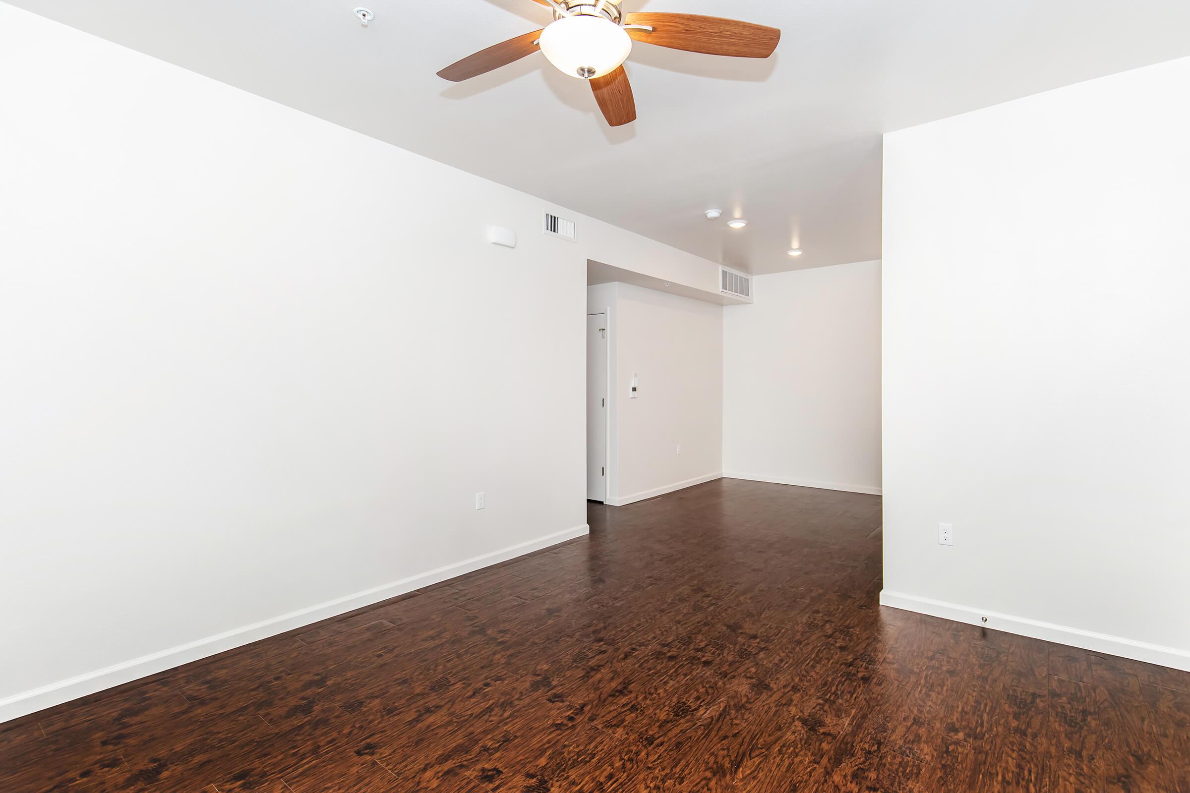 A spacious, unfurnished room with light-colored walls and a dark hardwood floor. A ceiling fan is visible, and there's a doorway leading to another room. The space is well-lit, with an open layout that emphasizes its size and potential for decoration or furnishing.