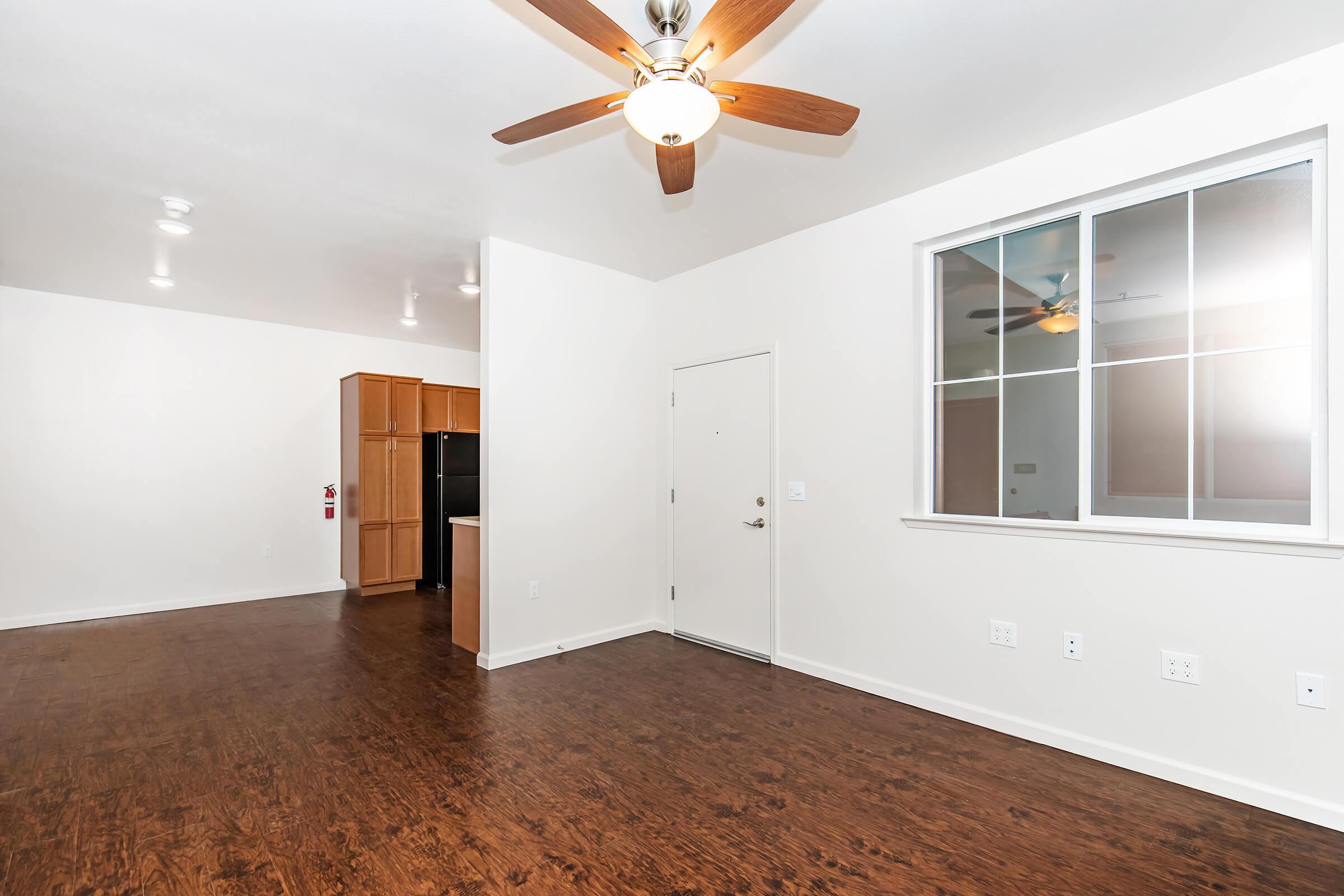 Interior view of a modern apartment featuring a spacious living area with dark wood flooring, a ceiling fan, and a large window. A doorway leads to the entrance, while a kitchenette with wooden cabinets and black appliances is visible in the background. The walls are painted in a light color for a bright atmosphere.