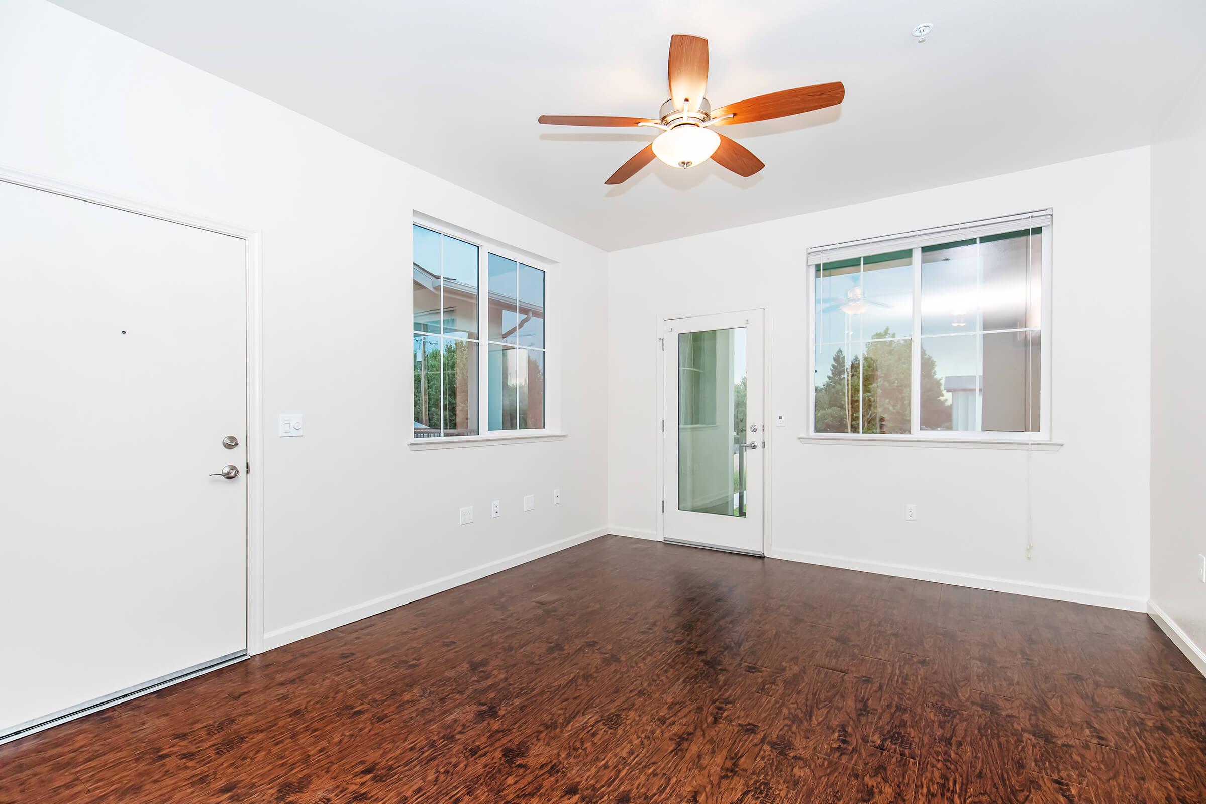 A spacious, unfinished room featuring light-colored walls, a ceiling fan, and two large windows. The flooring is dark wood, and there is a front door and a sliding glass door leading to a small outdoor area.
