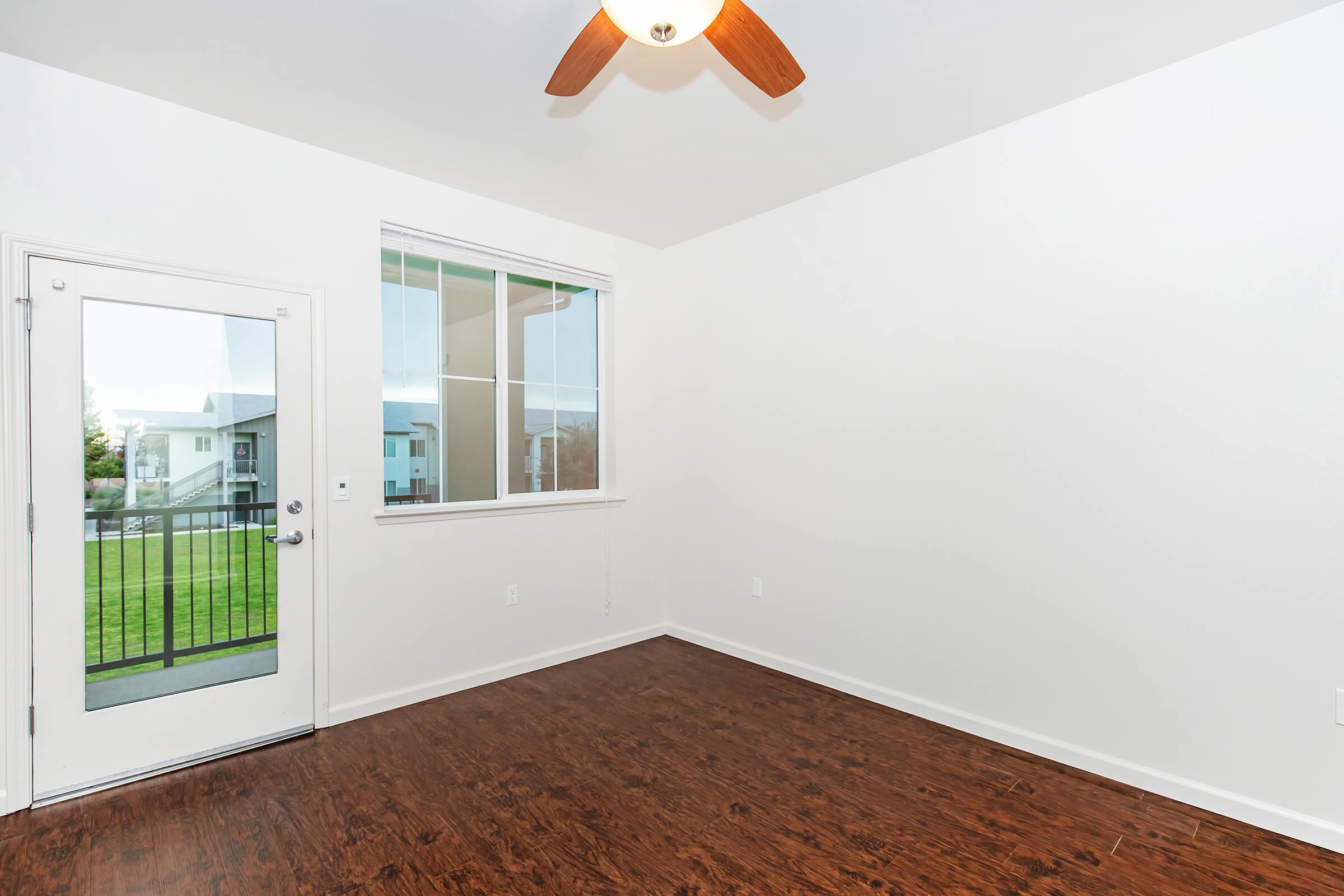 An empty room with a brown wooden floor, a ceiling fan with wooden blades, and a large window. A sliding glass door leads to an outdoor area with greenery visible outside. The walls are painted white, creating a bright and airy atmosphere.
