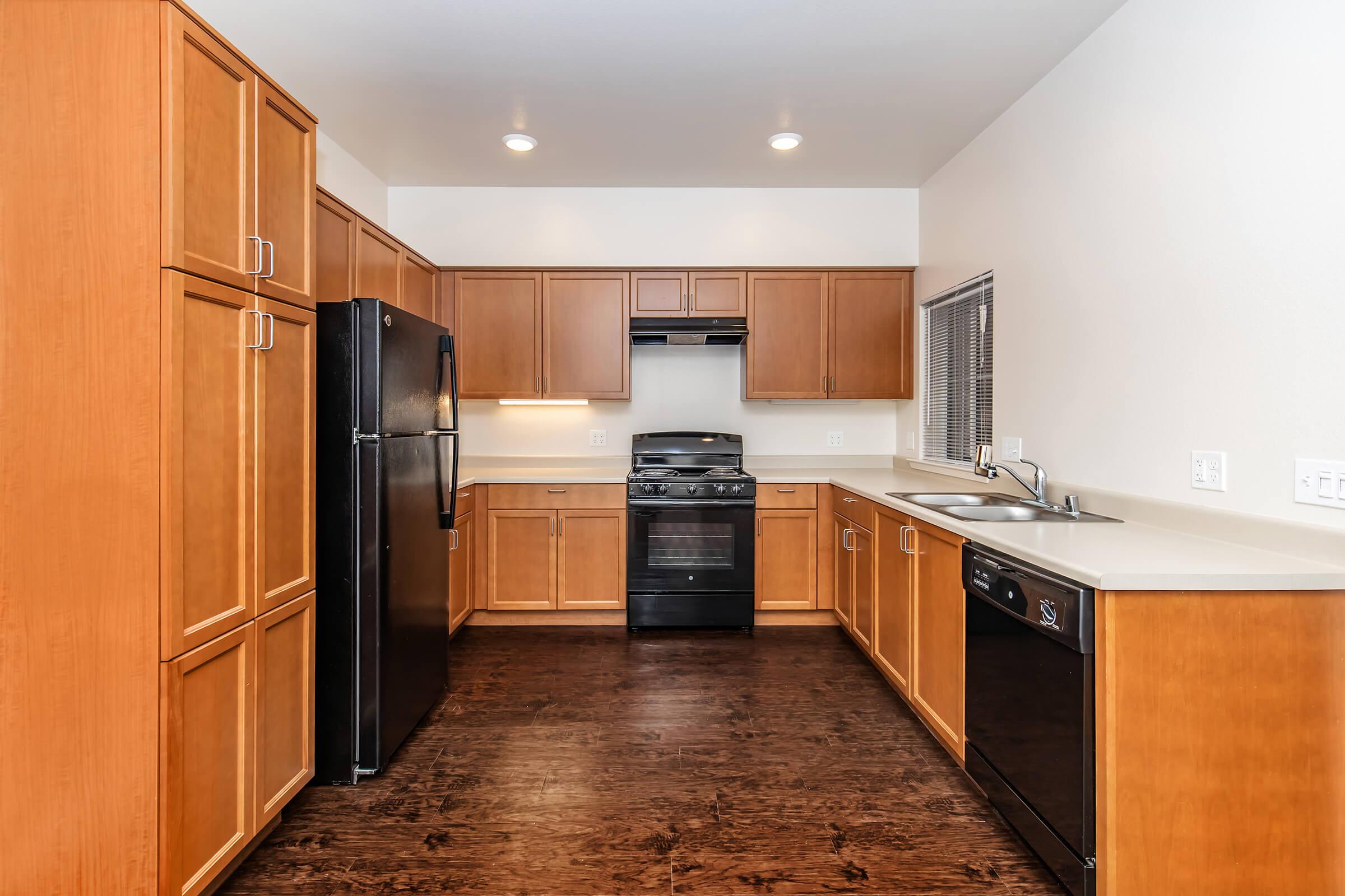Modern kitchen featuring wooden cabinetry, a black refrigerator, a black oven with a range, a stainless steel sink, and a dishwasher. The space has a light wall color and dark hardwood flooring, with natural light coming through a window.