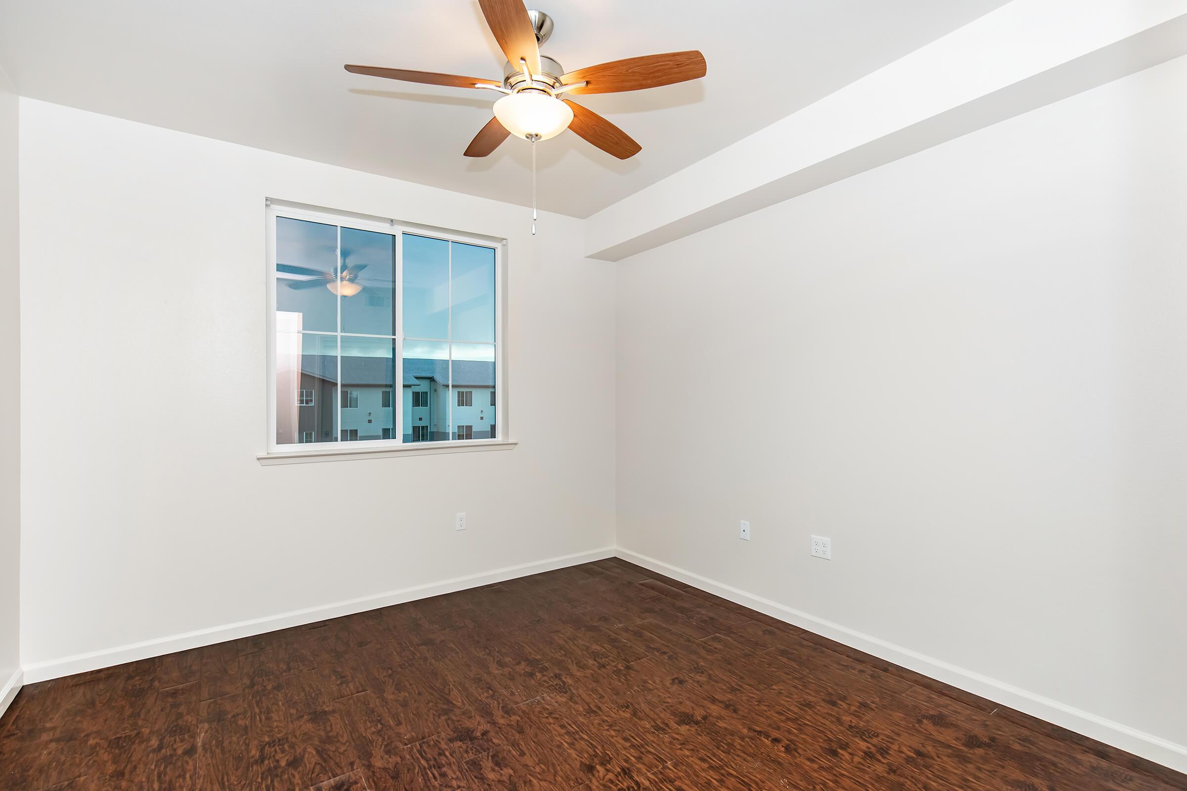 Empty room featuring a ceiling fan with wooden blades, light-colored walls, and a large window with a view. The floor is dark wood laminate, and there are no furnishings. The overall atmosphere is bright and clean, creating a spacious feel.