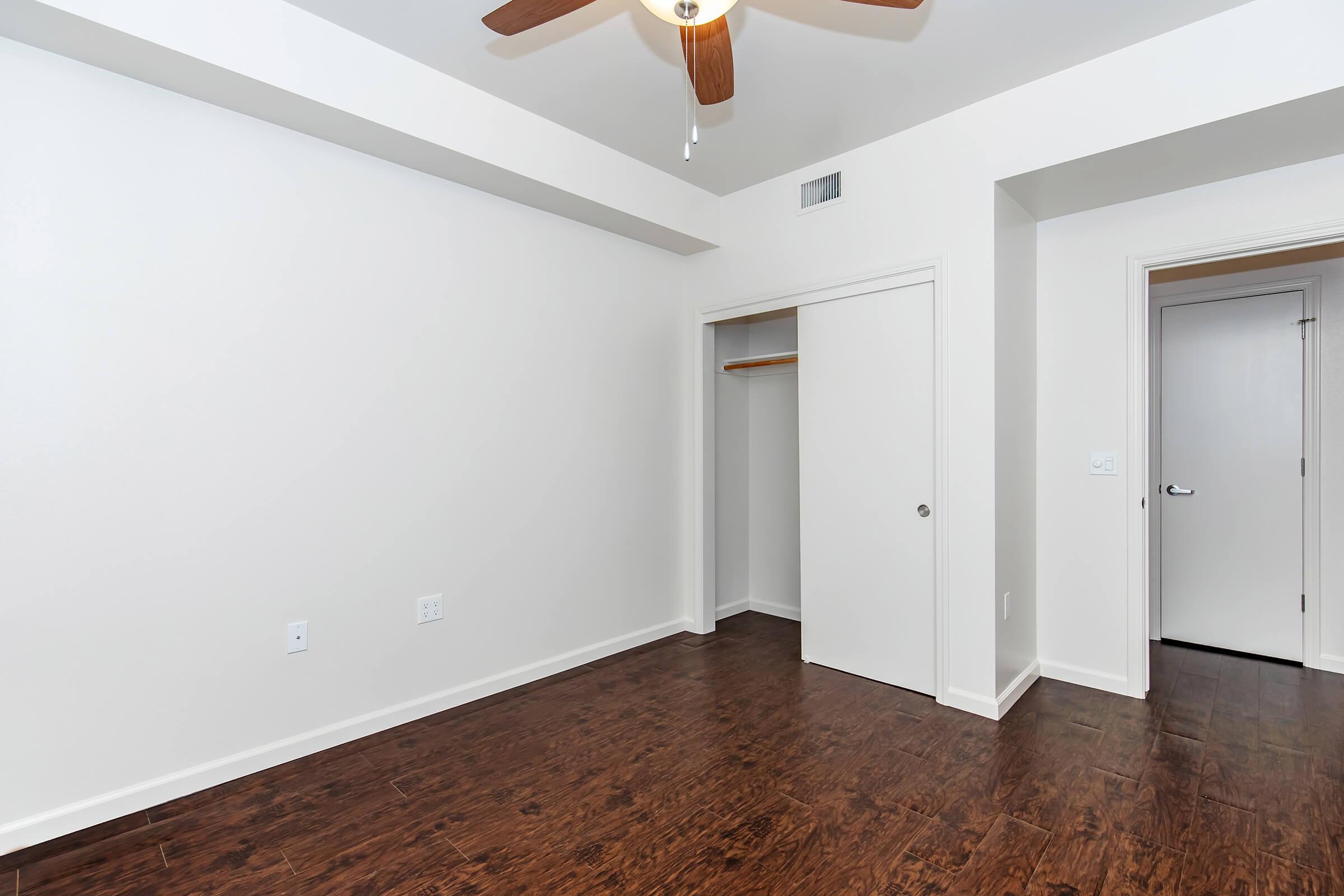 Empty interior room featuring light-colored walls and a dark wood floor. A ceiling fan is installed, and there is a closet with sliding doors on one side. A doorway leads to another space, enhancing the room's layout and lighting.