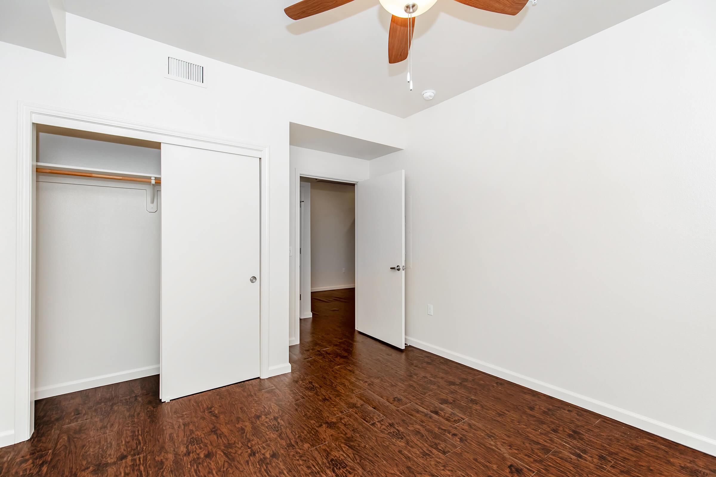 Empty room with a light-colored wall and dark wood flooring. A ceiling fan is visible, and there are open doors leading to a closet and another room. The space appears well-lit and clean, suitable for interior decorating or furnishing.