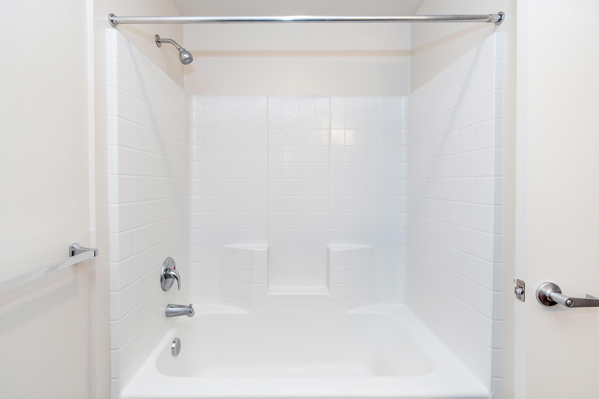 An empty, clean bathroom featuring a white tiled shower and bathtub. The walls are smooth and white, with a chrome showerhead and a shower curtain rail. There are two built-in shelves on either side of the bathtub, and a towel bar is visible on the left. The overall appearance is bright and minimalistic.