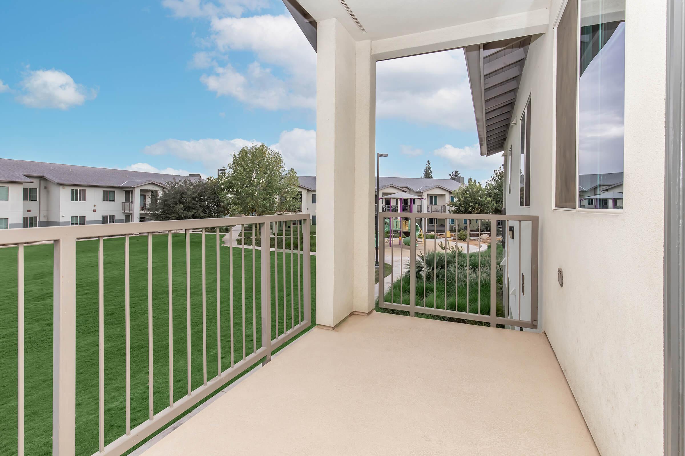 A view from a balcony overlooking a green lawn and nearby buildings. The space is bright with a clear blue sky and has a modern railing design. The surrounding area includes landscaped gardens, adding to the overall peaceful atmosphere.