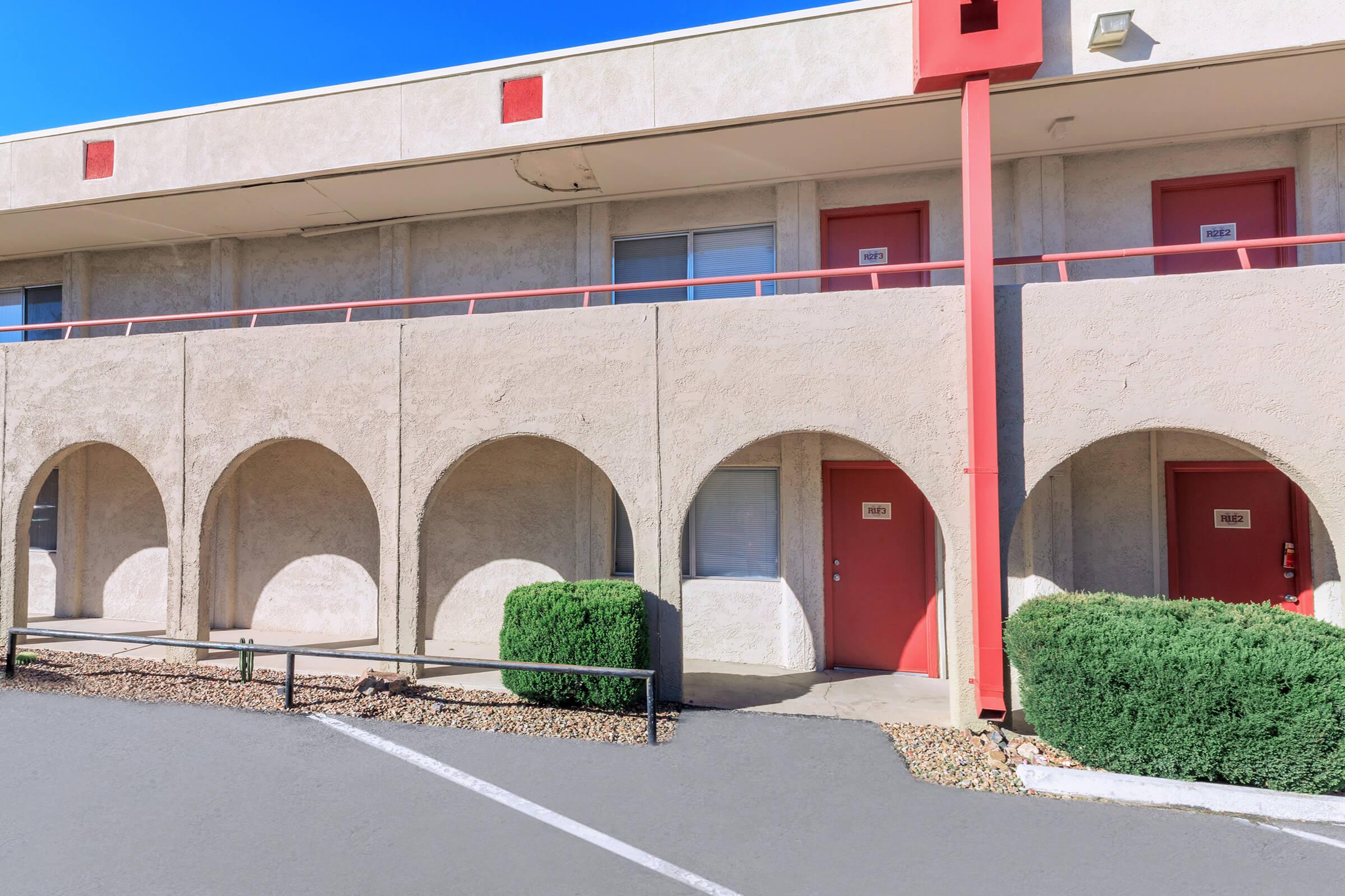 Exterior view of a two-story motel with arched entrances. The building features pale walls with a red accent, and there are several doors marked with room numbers. Shrubbery and a parking area are visible in the foreground, with a clear blue sky above.