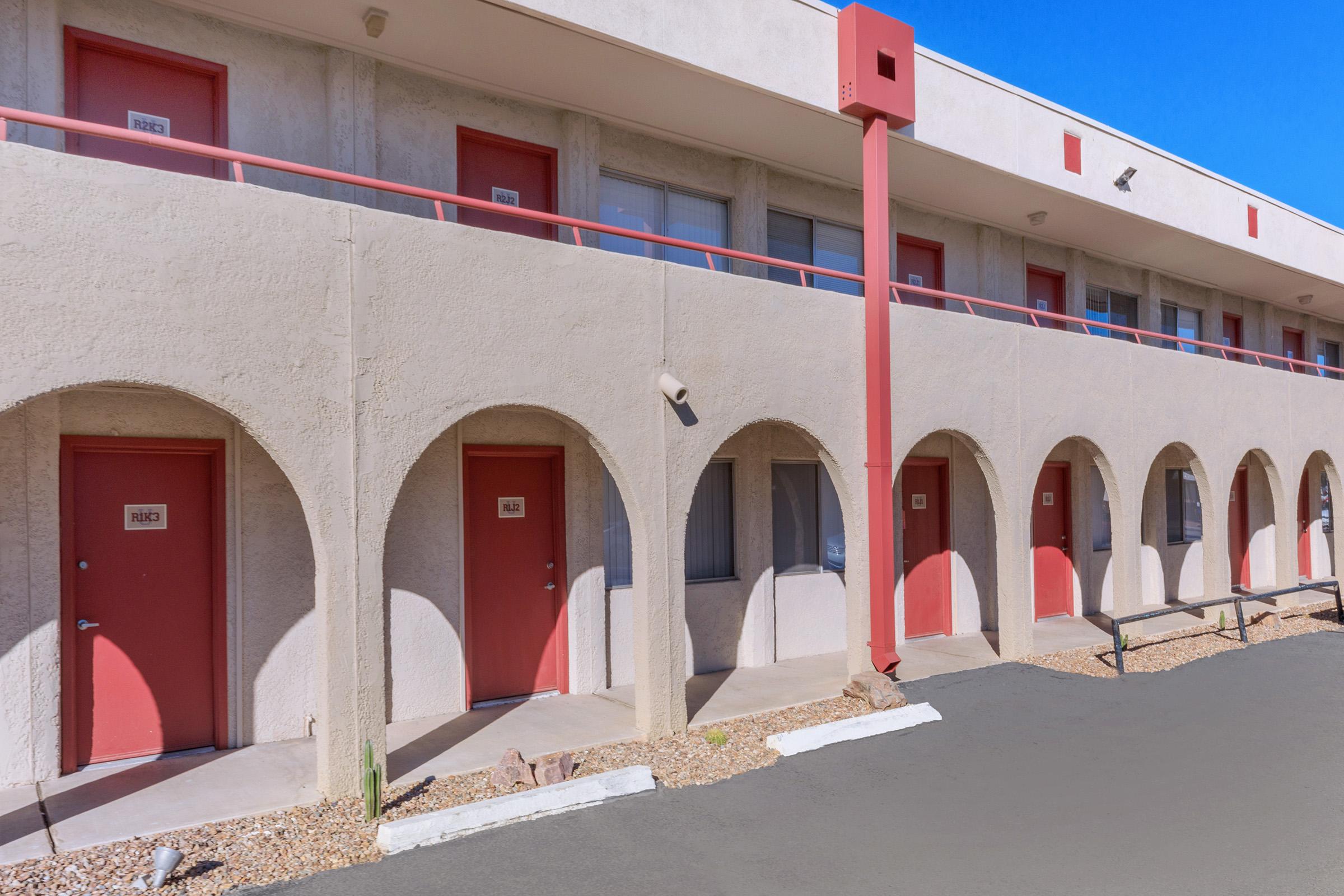 A row of motel units with red doors and decorative arches, set against a clear blue sky. The building features a pale exterior with a straight walkway and small gravel landscaping, highlighting the entrance of each room.