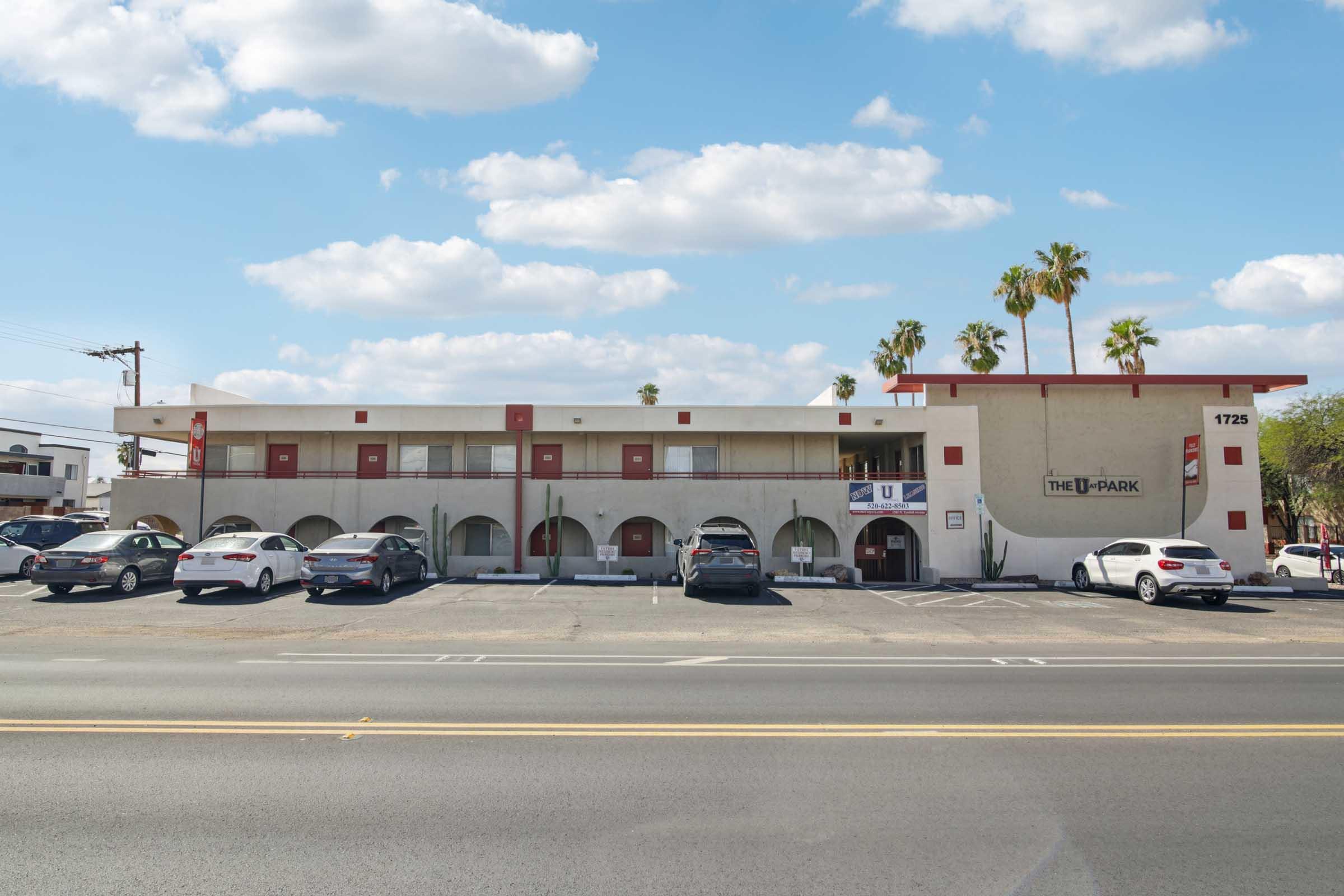 View of The U-Park motel, featuring a two-story building with red accents, palm trees, and a parking lot filled with cars. The sky is partly cloudy, and the structure has a sign displaying the name of the motel.
